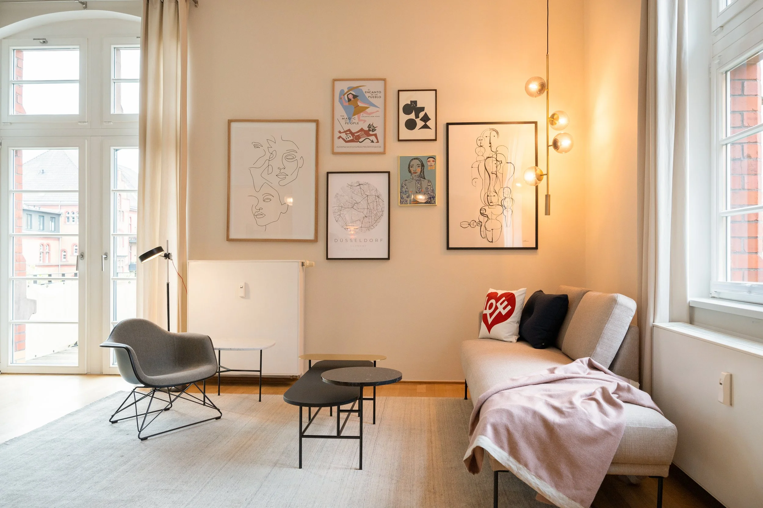 Helles Wohnzimmer mit großer Fensterfront, modernen Sitzmöbeln, einer dekorativen Bildergalerie an der Wand und warmem Licht.