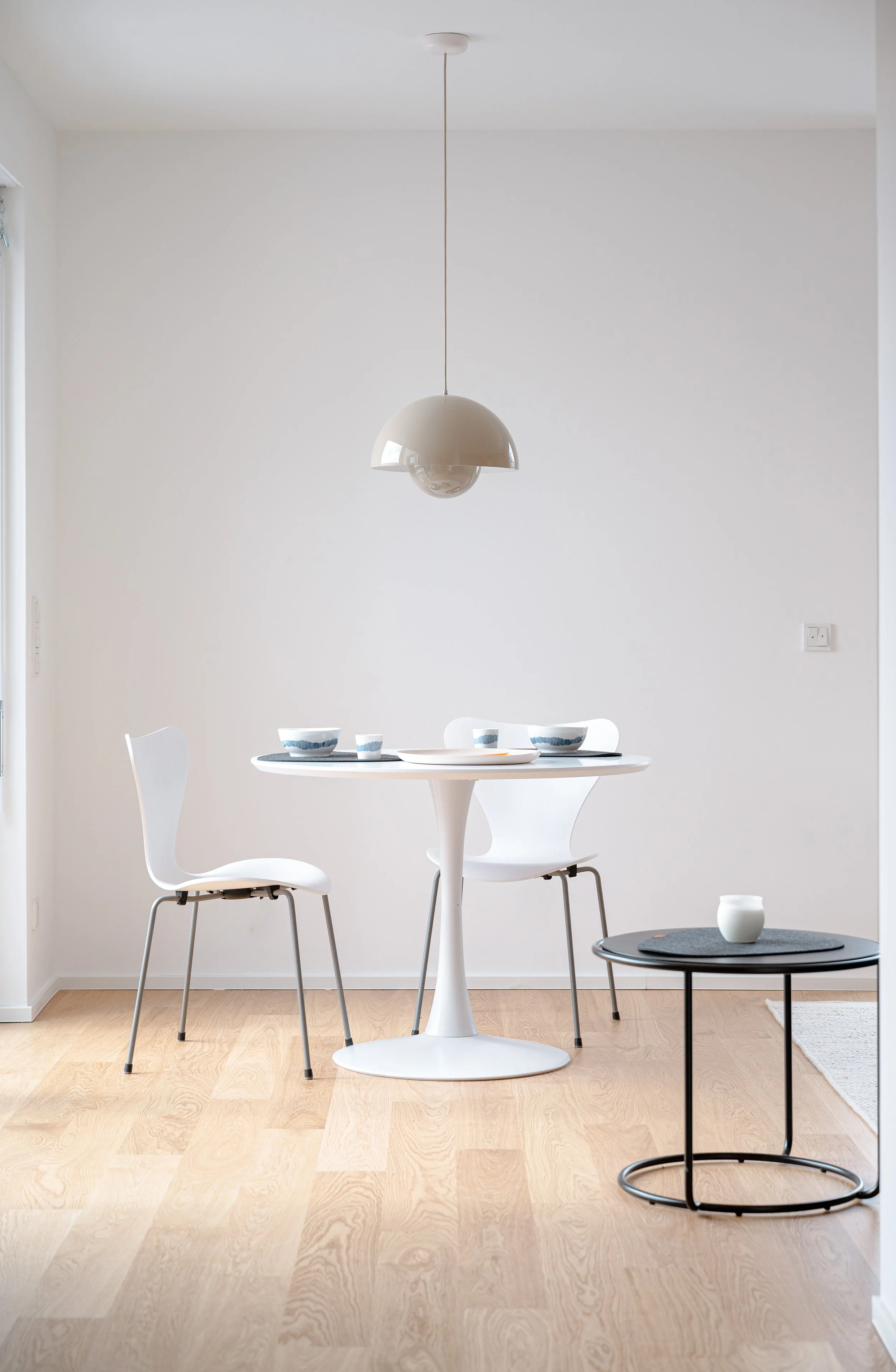 Ein minimalistisch eingerichteter Essbereich mit weißen Stühlen, einem weißen runden Tisch, einer schwarzen kleinen Beistelltisch mit einer weißen Vase, und einer modernen weißen Deckenleuchte, vor einer hellen weißen Wand und Holzboden.