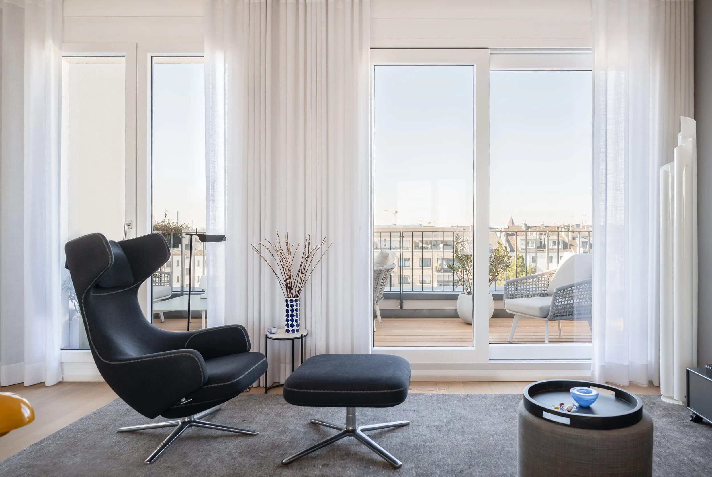 Wohnzimmer mit schwarzen Lounge-Stuhl, Couchtisch, Balkon mit Gartenmöbeln und urbidem Ausblick, weiße Vorhänge, natürliche Licht