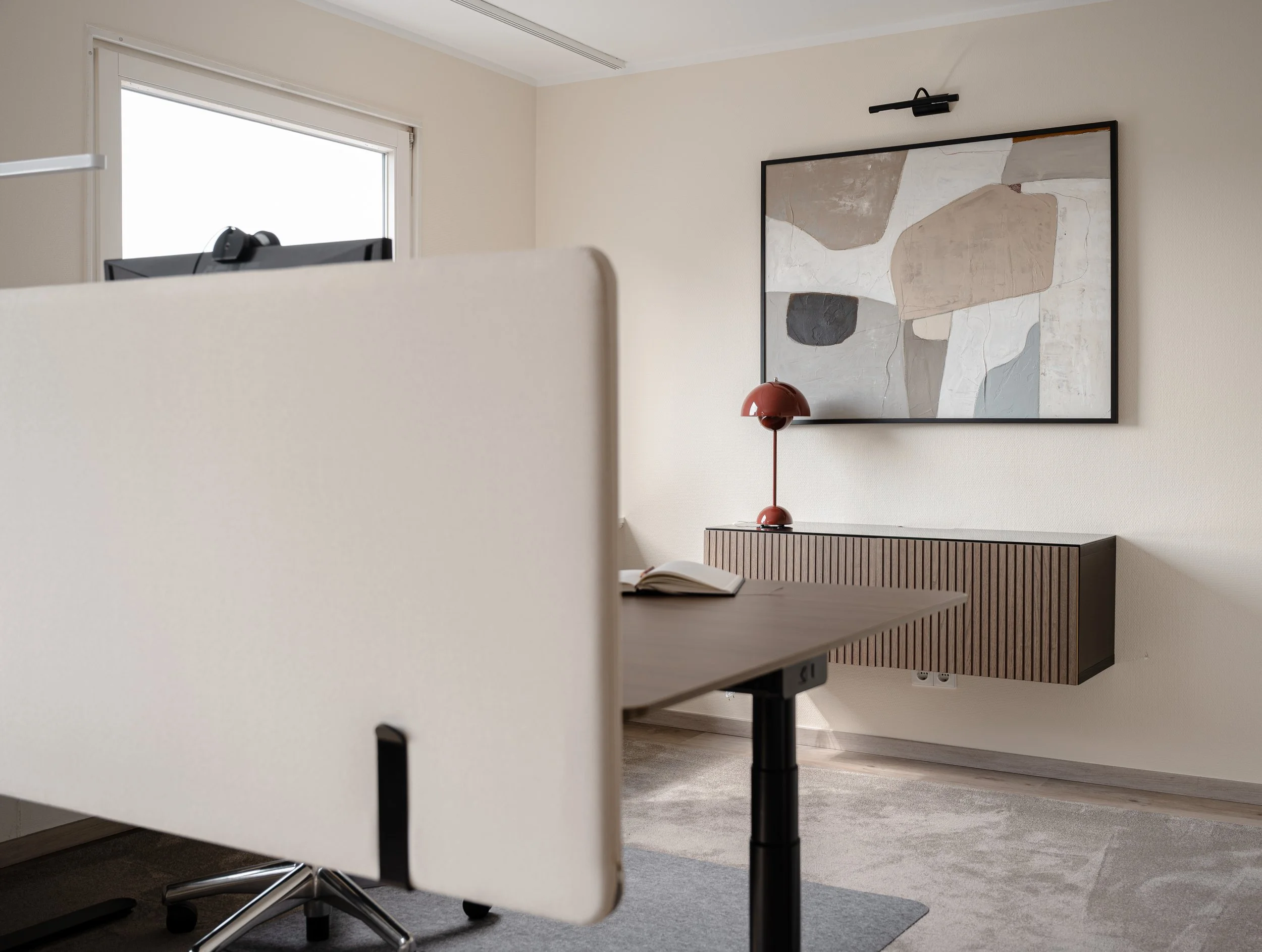 Moderner Arbeitsbereich mit einem weißen, rollbaren Raumteiler, einem Schreibtisch mit offenen Buch, einem abstrakten Gemälde an der Wand, einer roten Tischlampe auf einem Holz-Schrank und einem Fenster im Hintergrund.