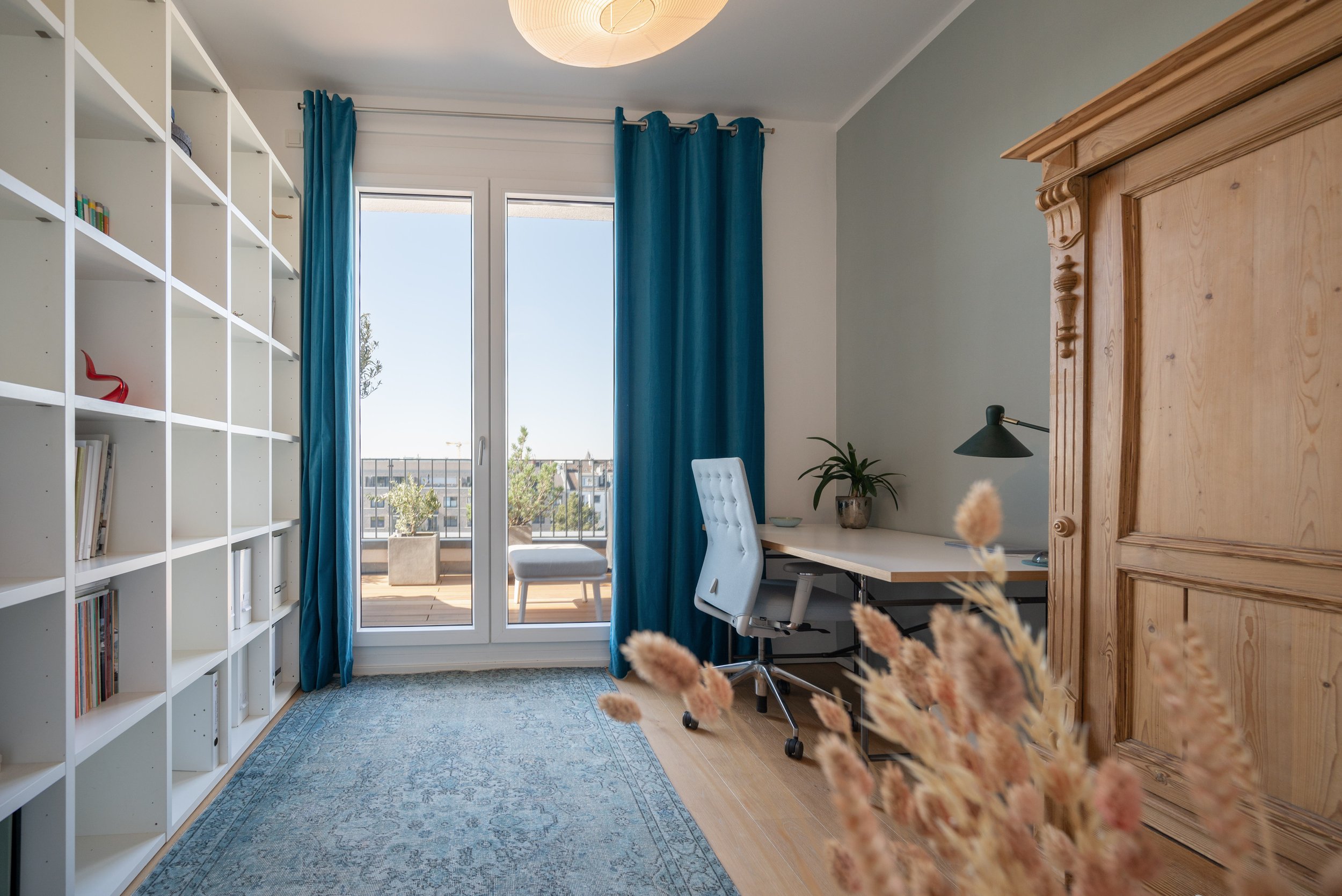 Ein Arbeitszimmer mit einem großen Bücherregal auf der linken Seite, einem Schreibtisch mit einem weißen Bürostuhl vor einem Fenster mit blauen Vorhängen, einem kleinen Balkon draußen, einer Grünen Pflanze und einem braunen Schrank auf der rechten Se