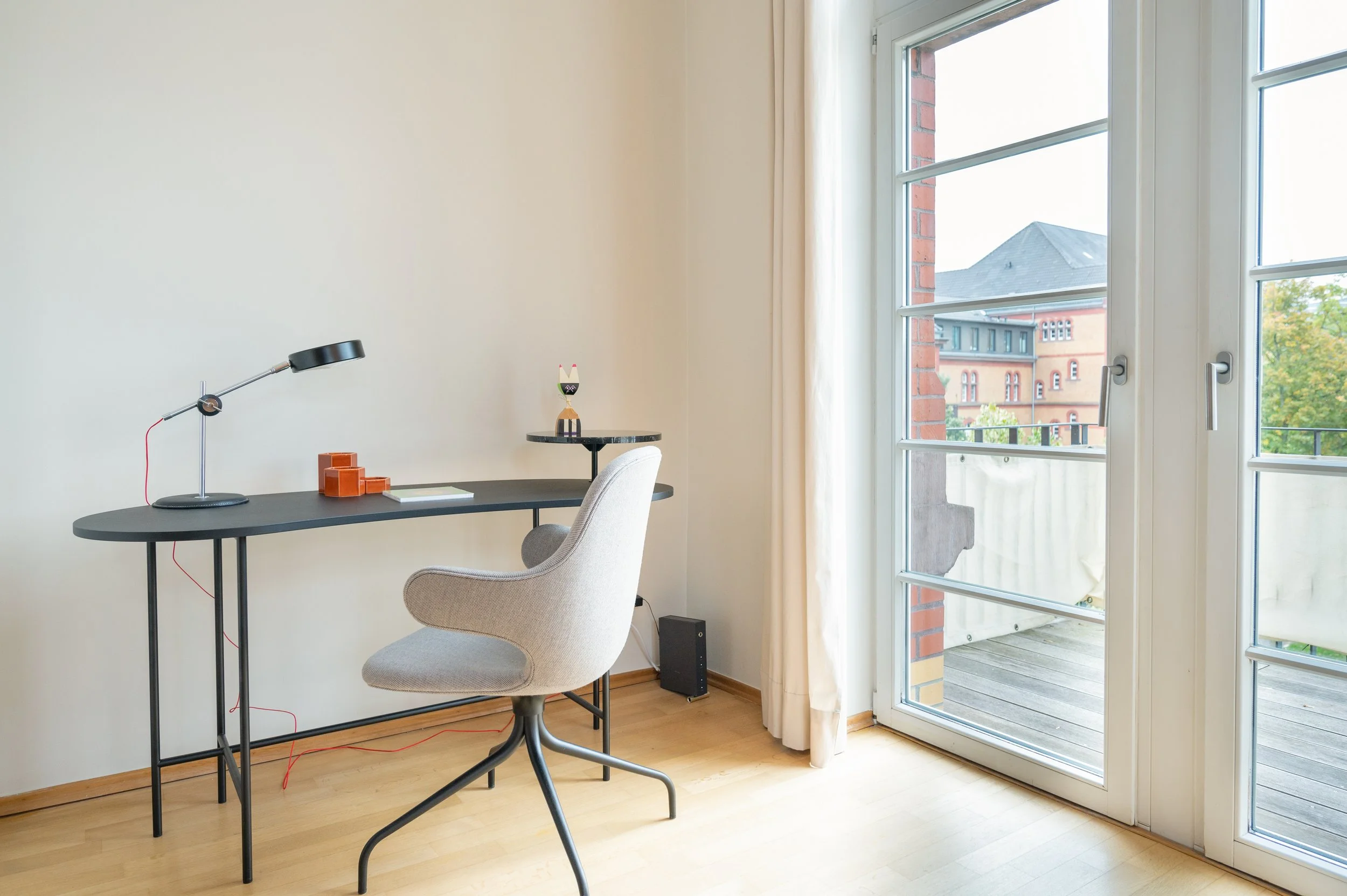 Ein minimalistischer Arbeitsbereich mit einem schwarzen Schreibtisch, einem weißen Bürostuhl, einer schwarzen Tischlampe, einigen kapla Steinen, einer kleinen Figur und einem kleinen schwarzen Lautsprecher. Es gibt große Fenster mit Blick auf Nachbar