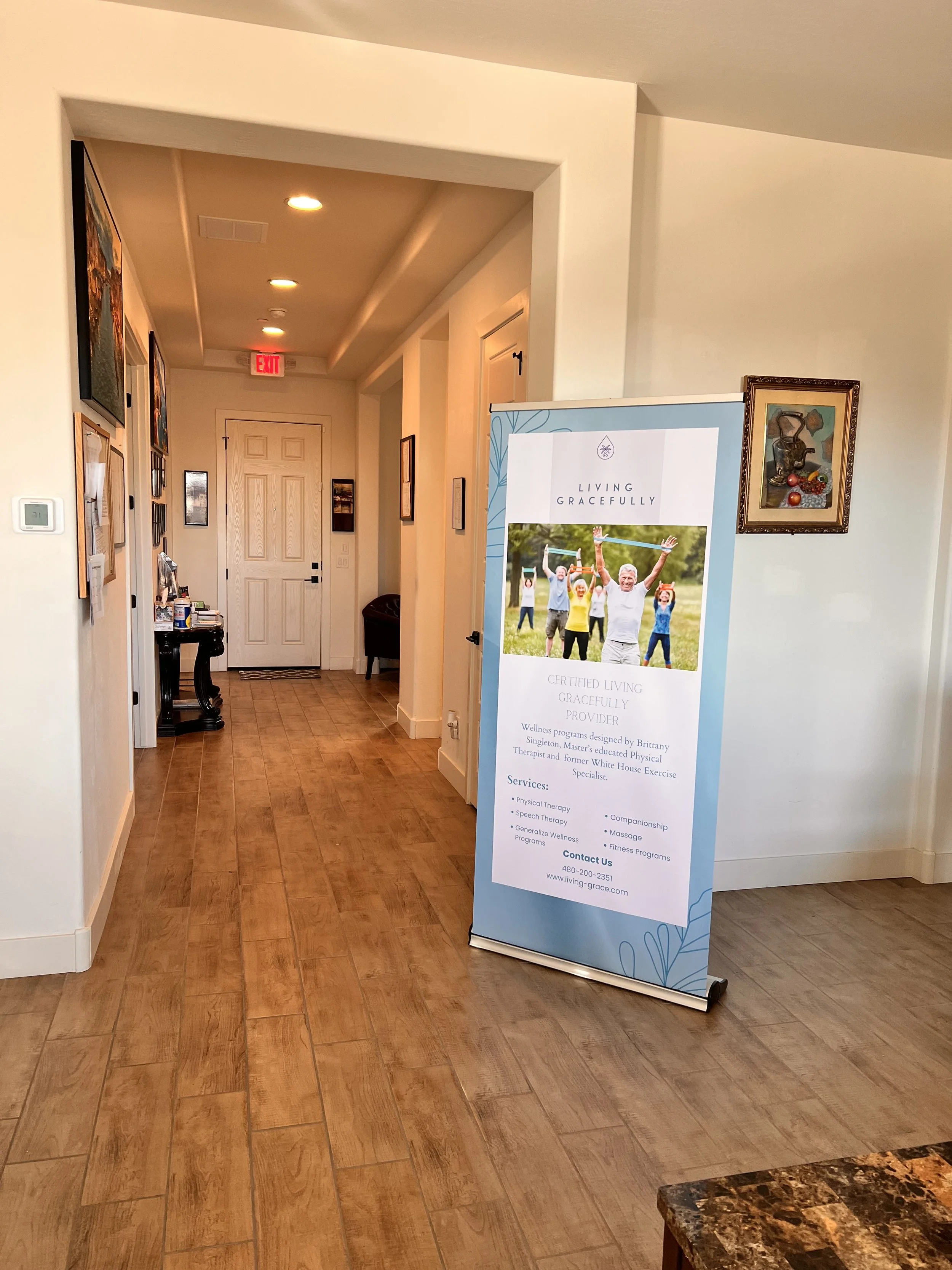 Interior of a wellness center lobby with a promotional sign for Living Gracefully services, wooden flooring, and framed art on the walls.