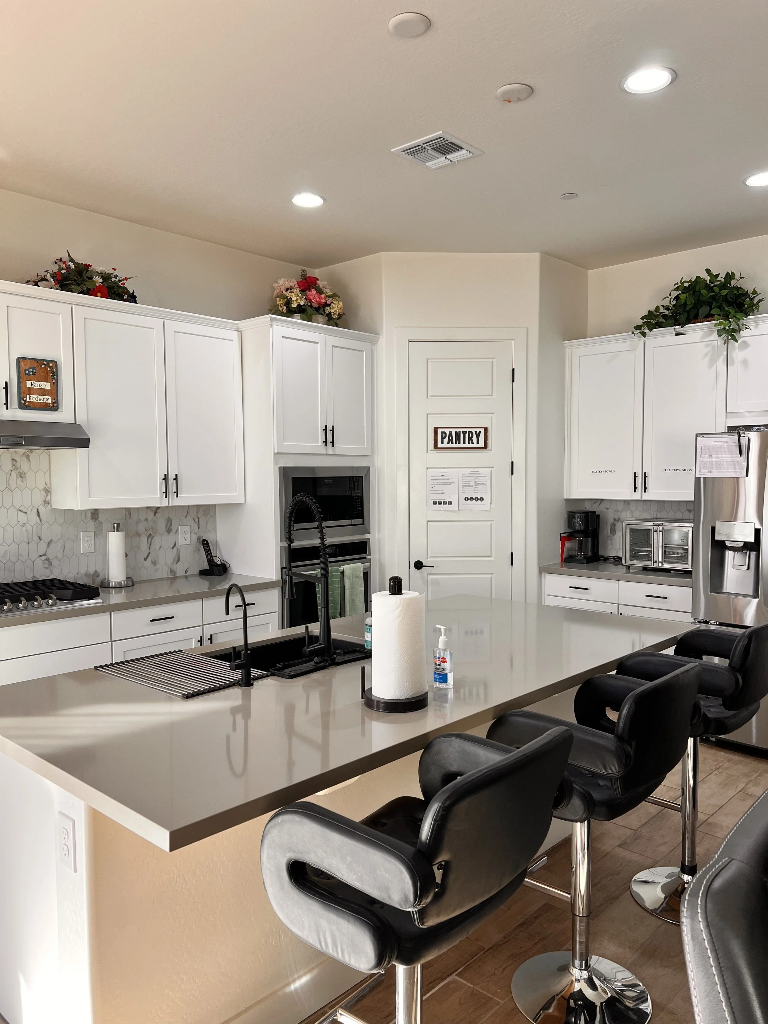 Modern kitchen with white cabinets, a large central island with black bar stools, stainless steel appliances, and decorative flowers on top of the cabinets.