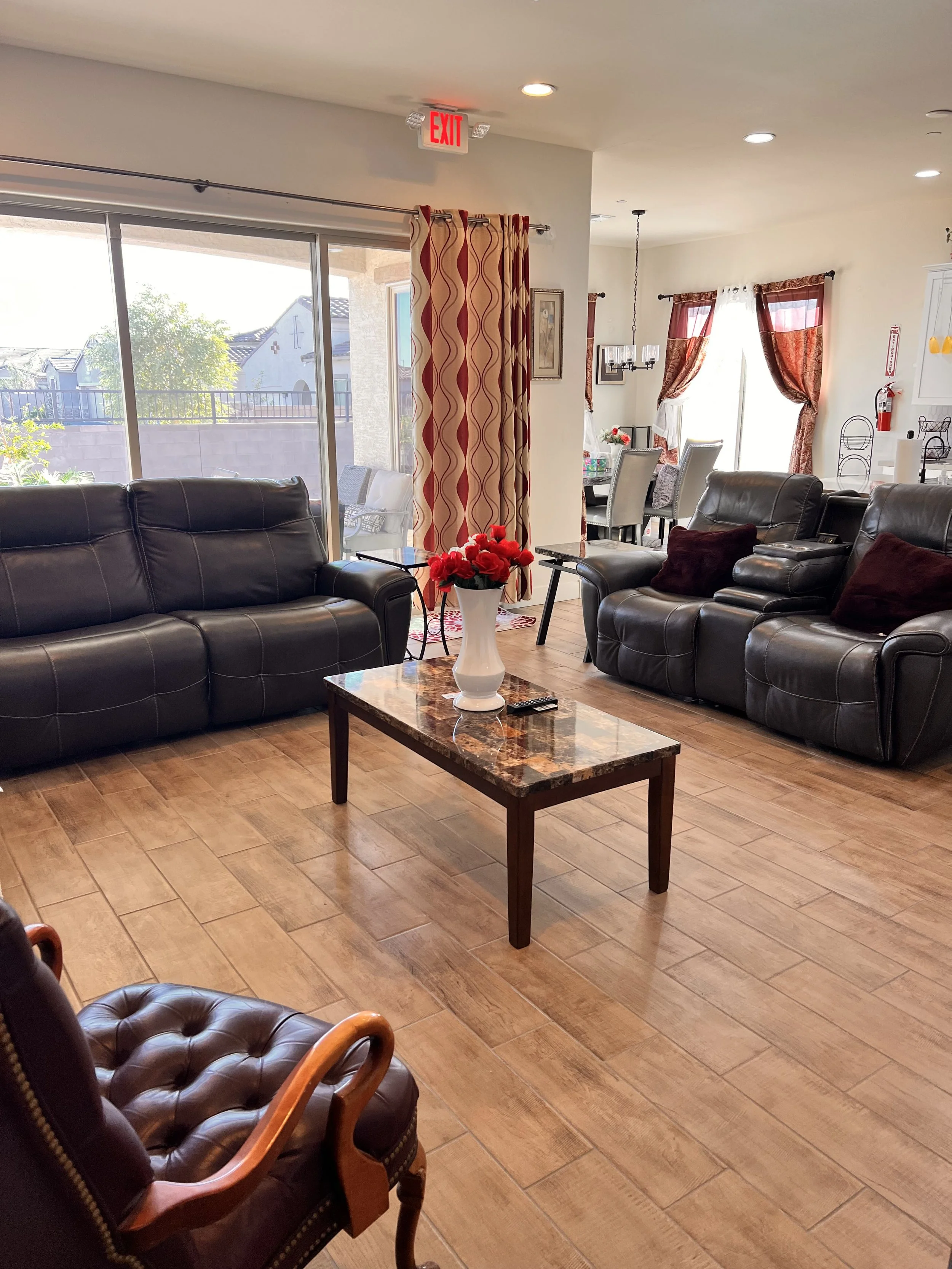 Living room with leather sofas, a glass sliding door with a striped curtain, wooden flooring, a table with a vase of red flowers, a dining area in the background with curtains, and an exit sign on the ceiling.