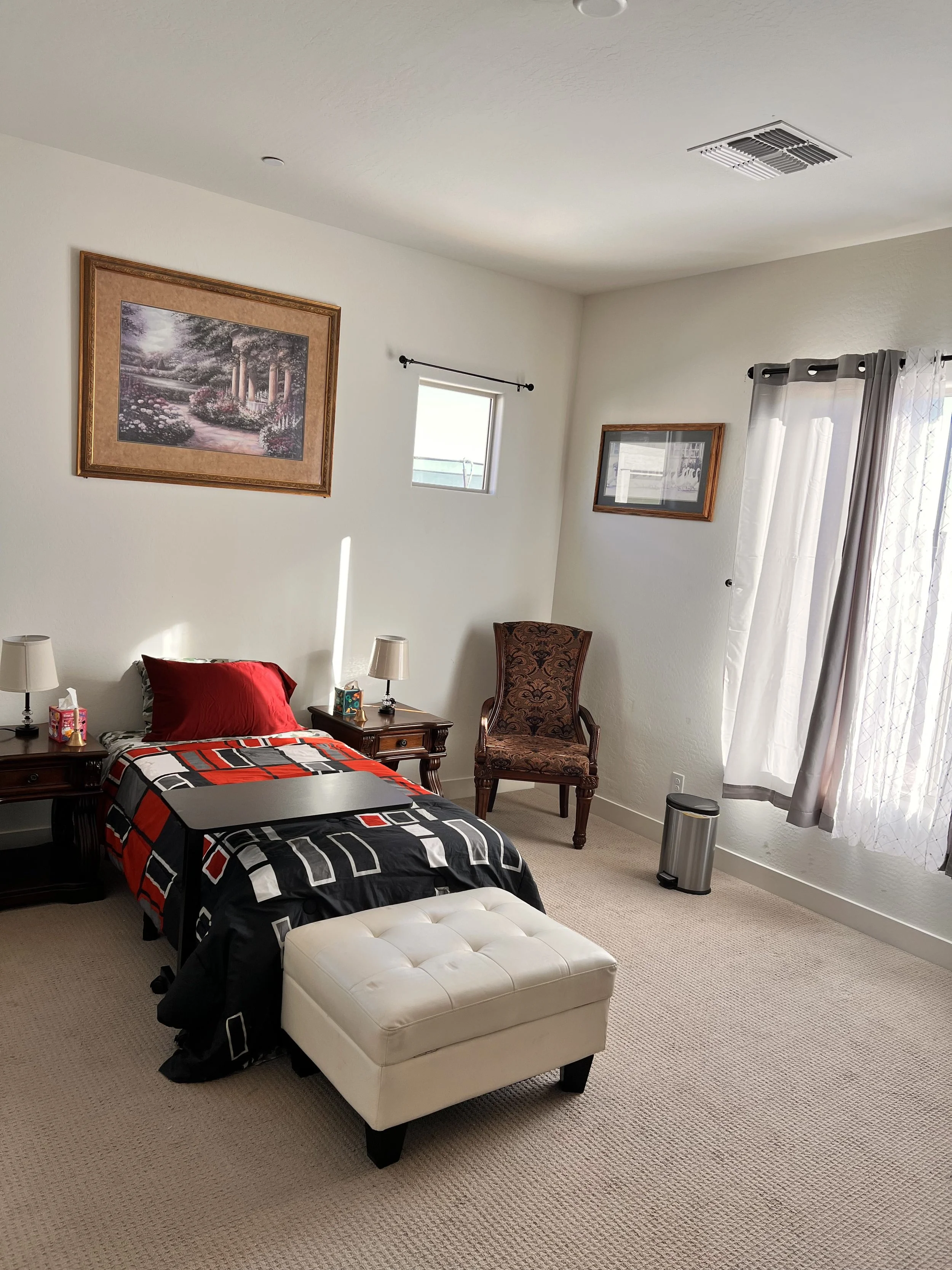 A bedroom with a single bed, red pillow, and patterned black comforter, with nightstands on both sides, each with a small lamp and tissue box. An upholstered chair and a white tufted ottoman are in view. There are framed pictures on the walls, a smal