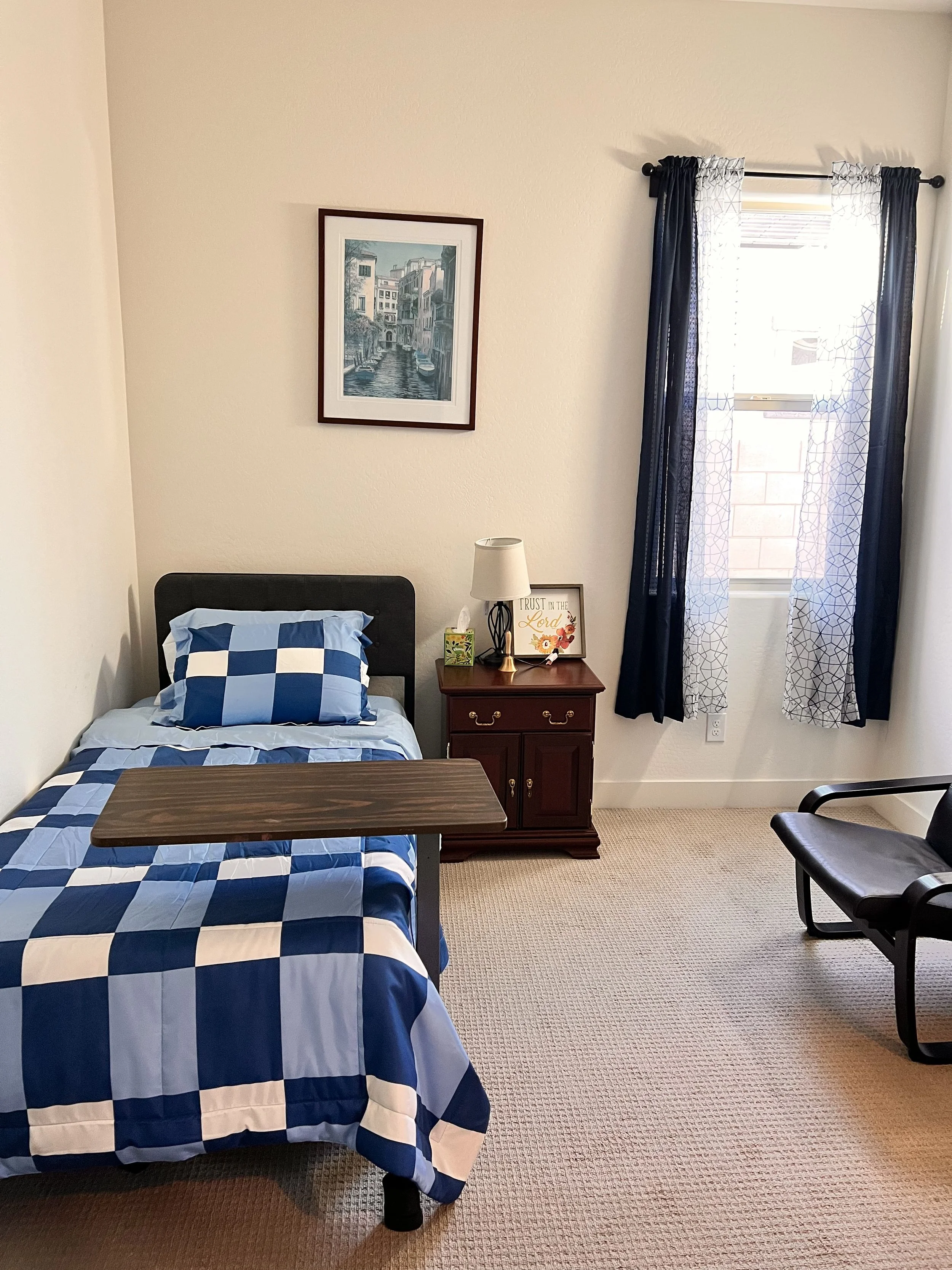 A bedroom with a single bed covered in a blue and white checkered comforter and pillow. A black headboard, bedside table with a lamp, framed artwork, and a framed sign. A window with navy blue and white curtains, and a black chair against the wall. T