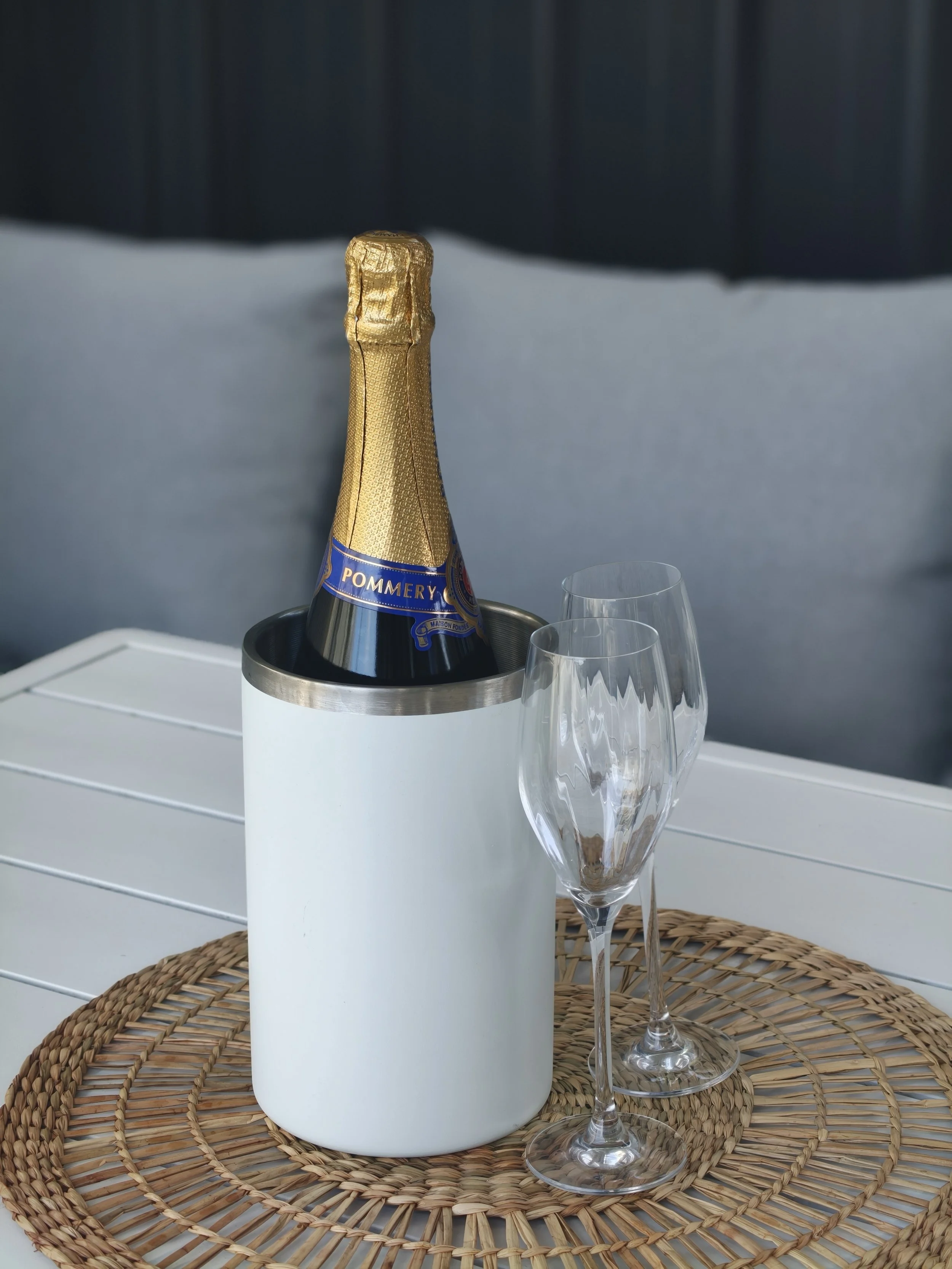 A bottle of Pommery champagne with gold foil, placed in a white ice bucket, on a woven placemat, with empty champagne glasses nearby, against a dark background.