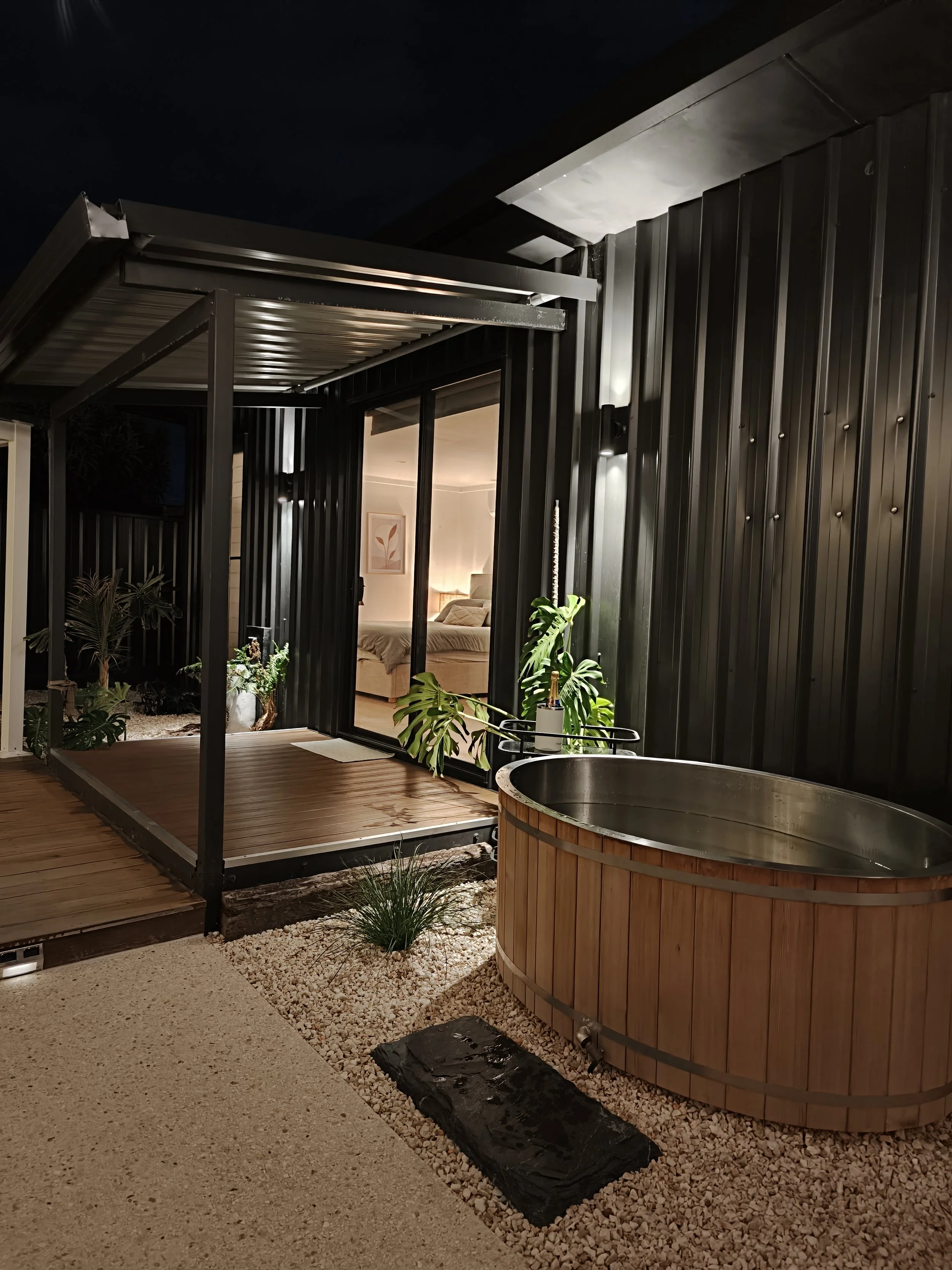 Nighttime view of a modern outdoor patio with a black metal roof, black metal panels, glass sliding doors, potted plants, a wooden hot tub, gravel, and artificial turf.