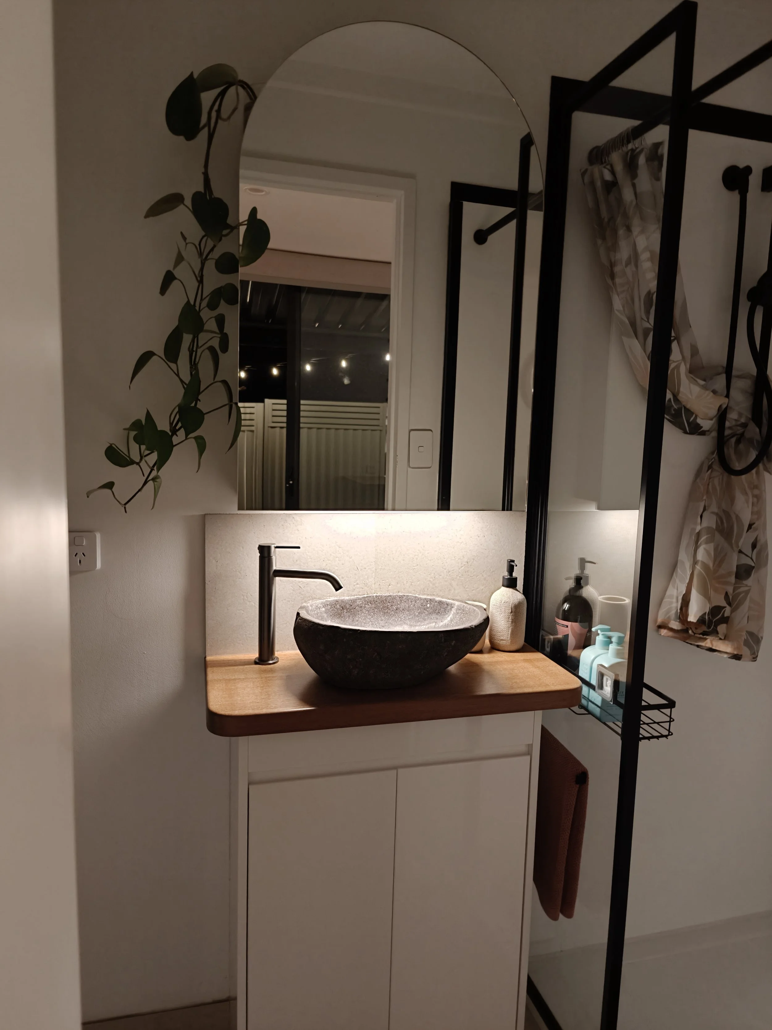 Bathroom vanity with a black stone sink, a wooden countertop, a modern black faucet, a mirror, and various toiletries. A plant is visible on the left side, and a window with patterned curtains is on the right.