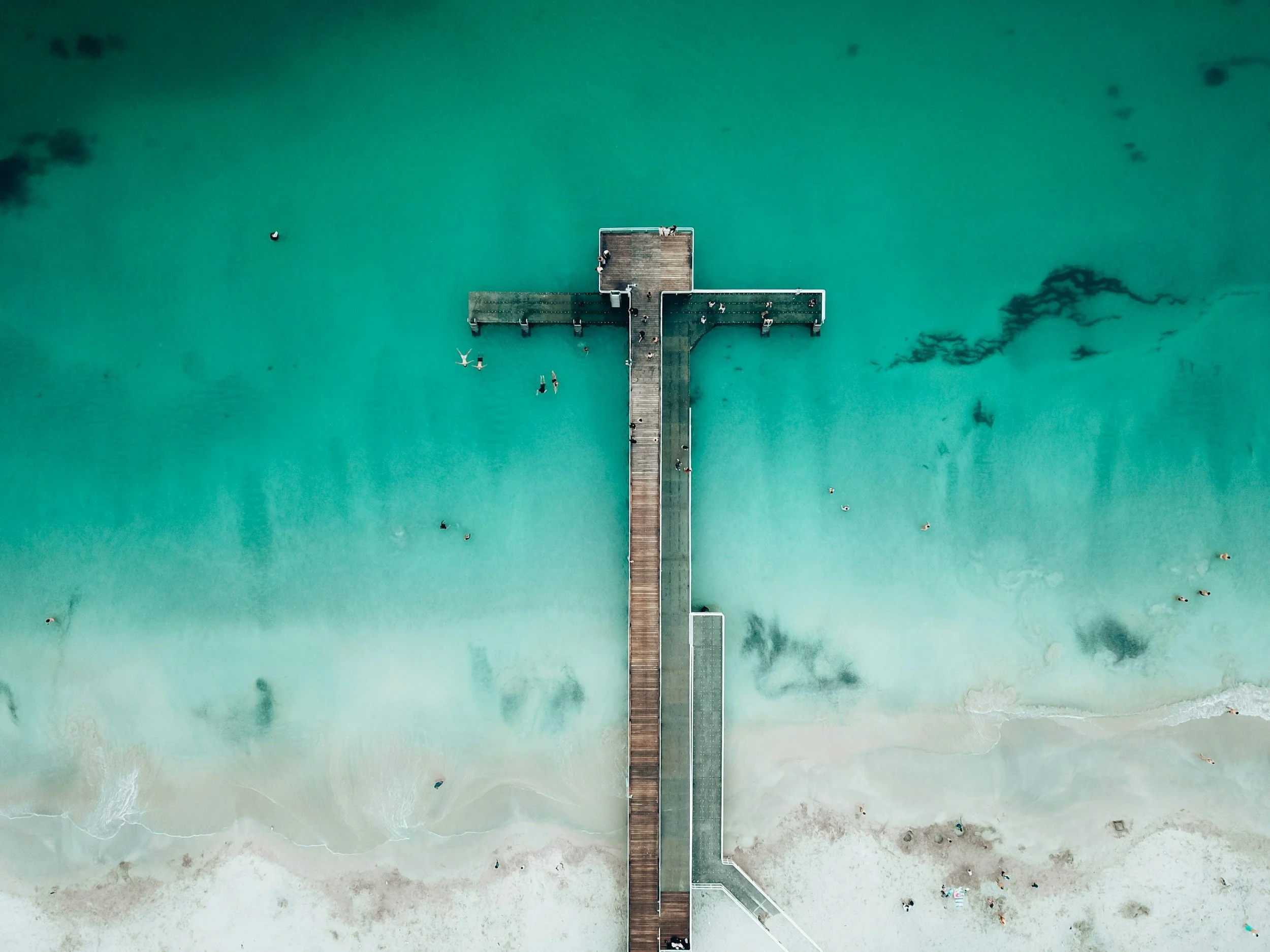 Aerial view of a wooden pier extending into clear teal ocean water with people swimming and relaxing nearby, sandy beach at the bottom.