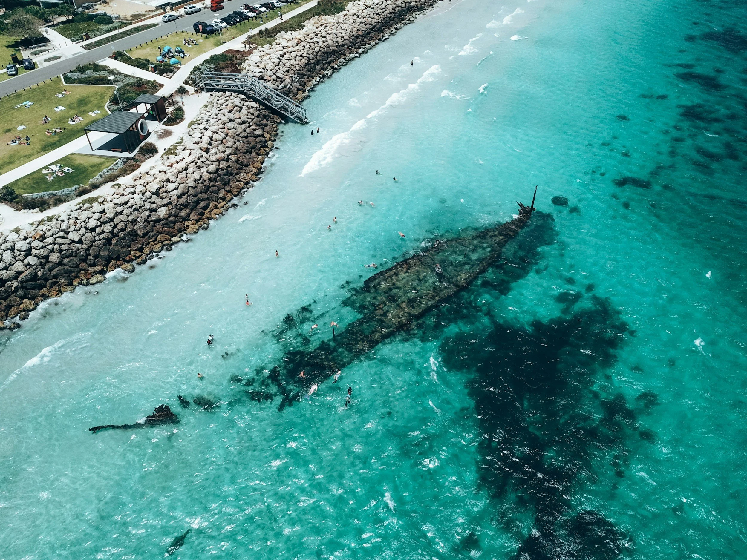 A shipwreck submerged in shallow turquoise waters near a rocky shoreline with people swimming and a park area with people relaxing on the grass.