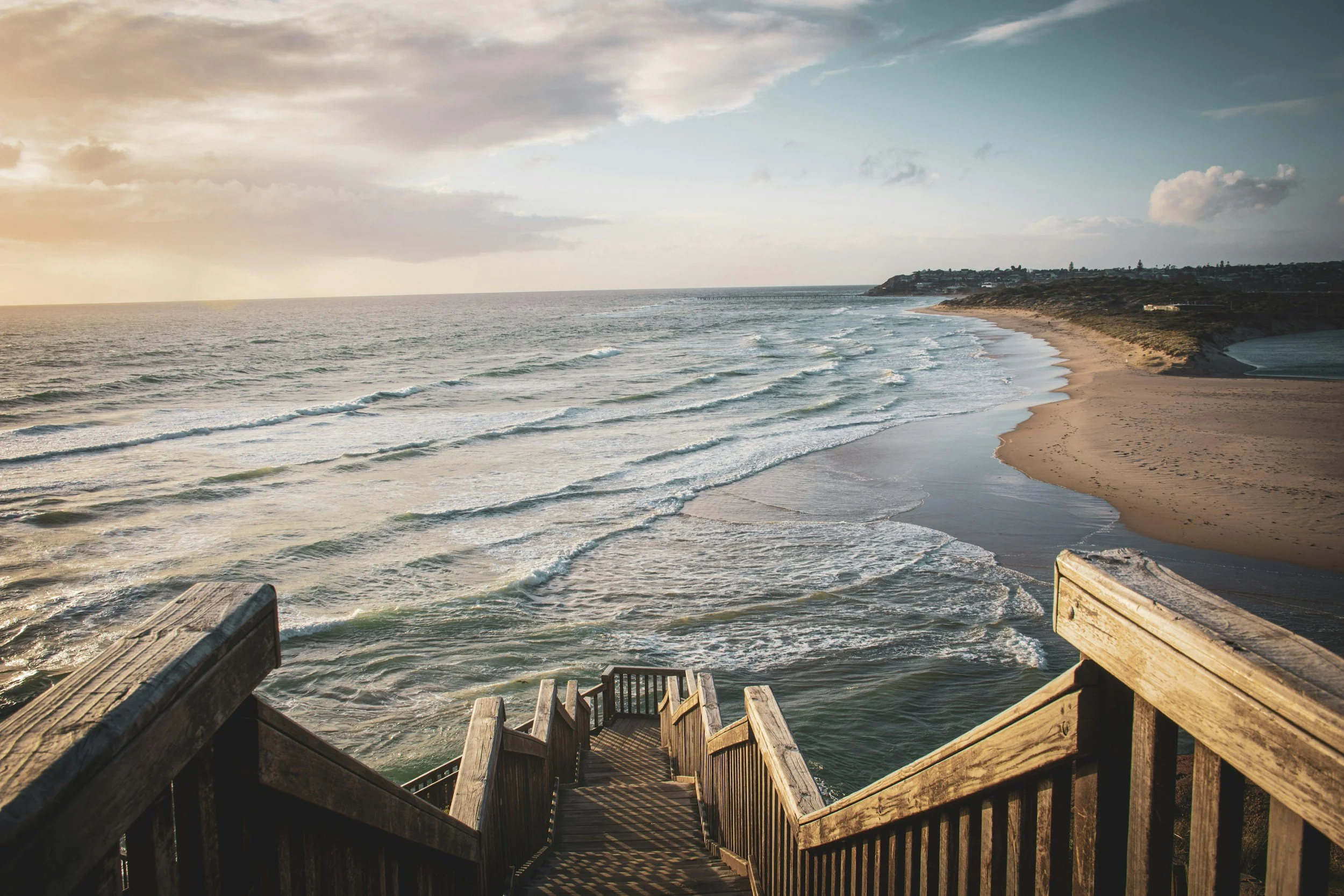 Wooden stairs leading down to a sandy beach with waves, overlooking the ocean and distant coastline at sunset.