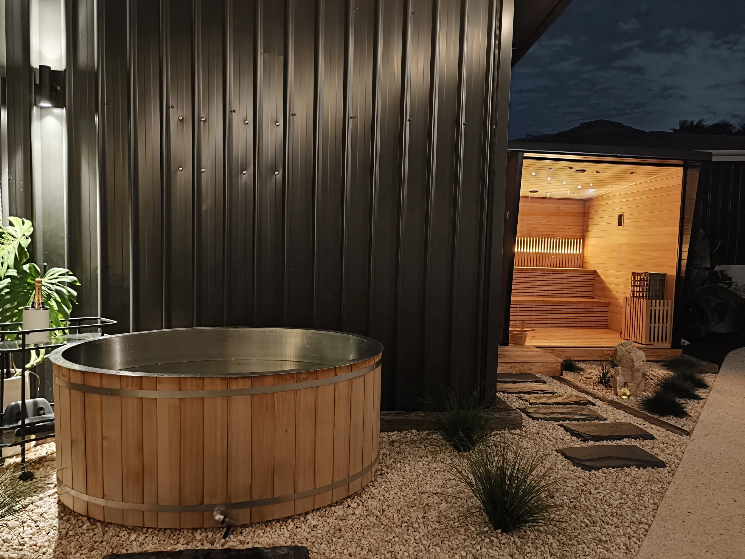 An outdoor nighttime scene featuring a modern enclosed sauna with warm lighting, a round hot tub with wooden paneling, and landscape with gravel, stones, and plants.