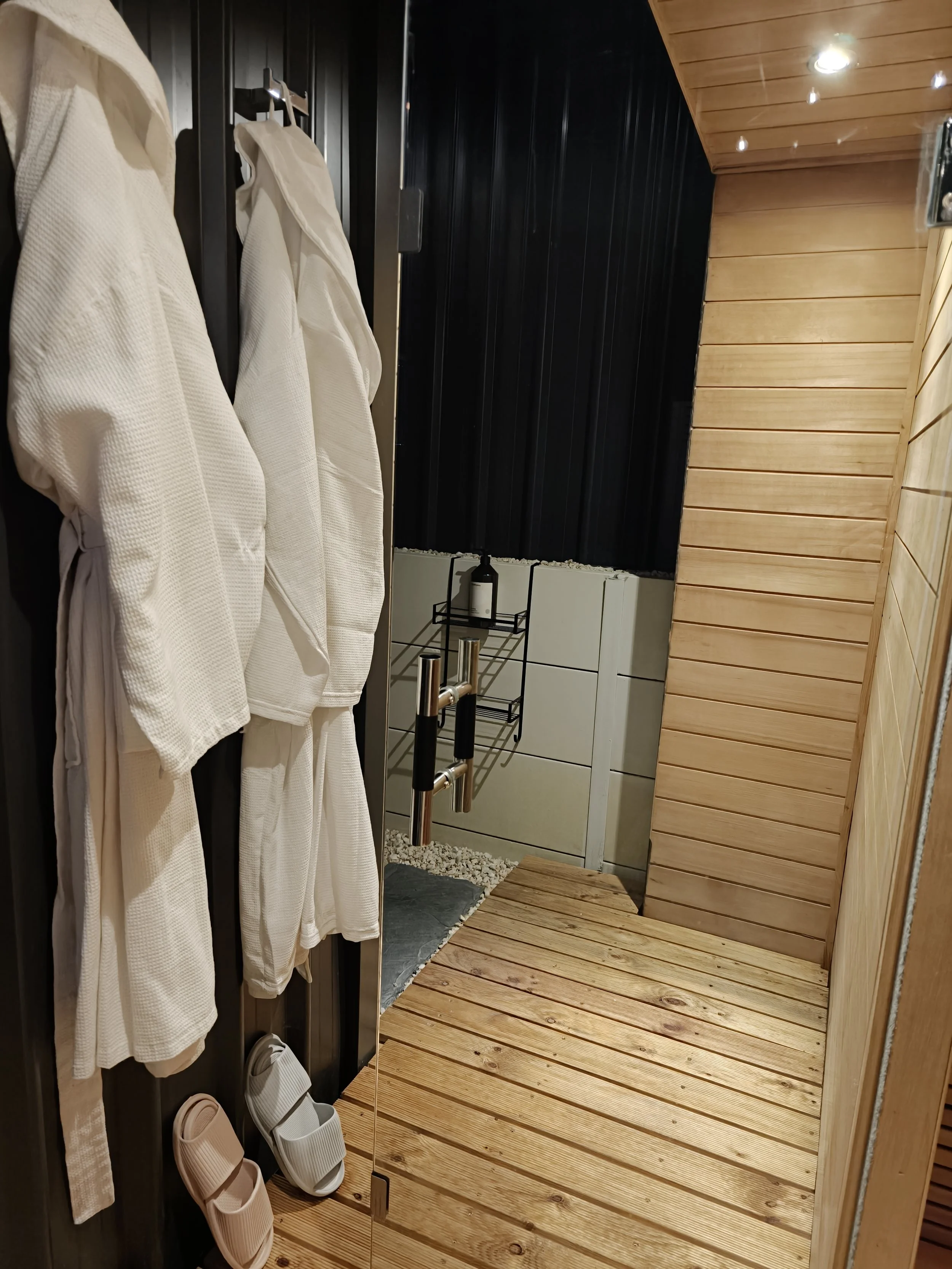 A sauna room with wooden flooring and walls, white bathrobes hanging on hooks, and slippers on the floor.
