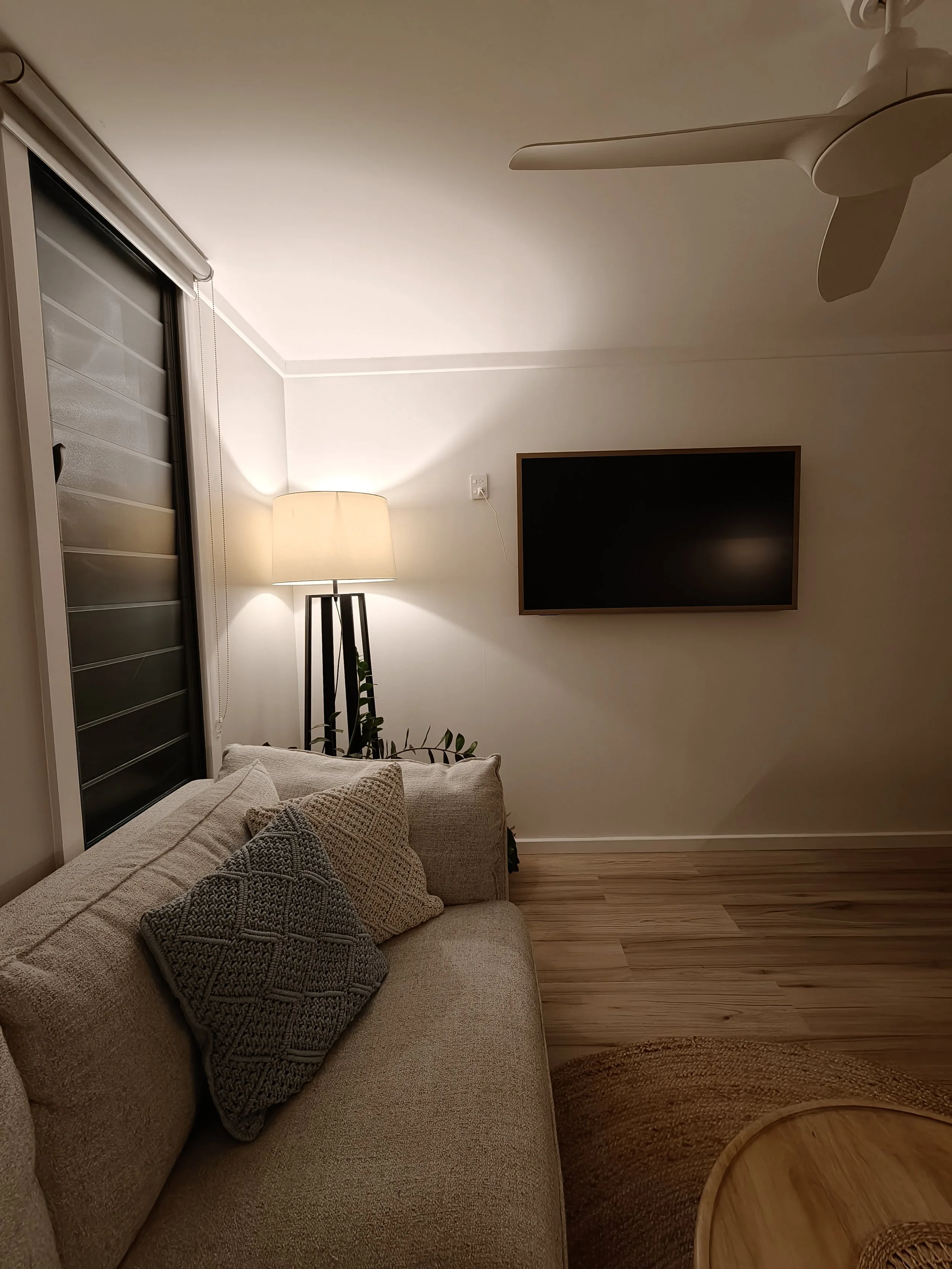 Living room with a beige sofa covered with knitted pillows, a floor lamp emitting warm light, a flat-screen TV on the wall, a sliding glass door with blinds, and a wooden side table with a basket underneath.