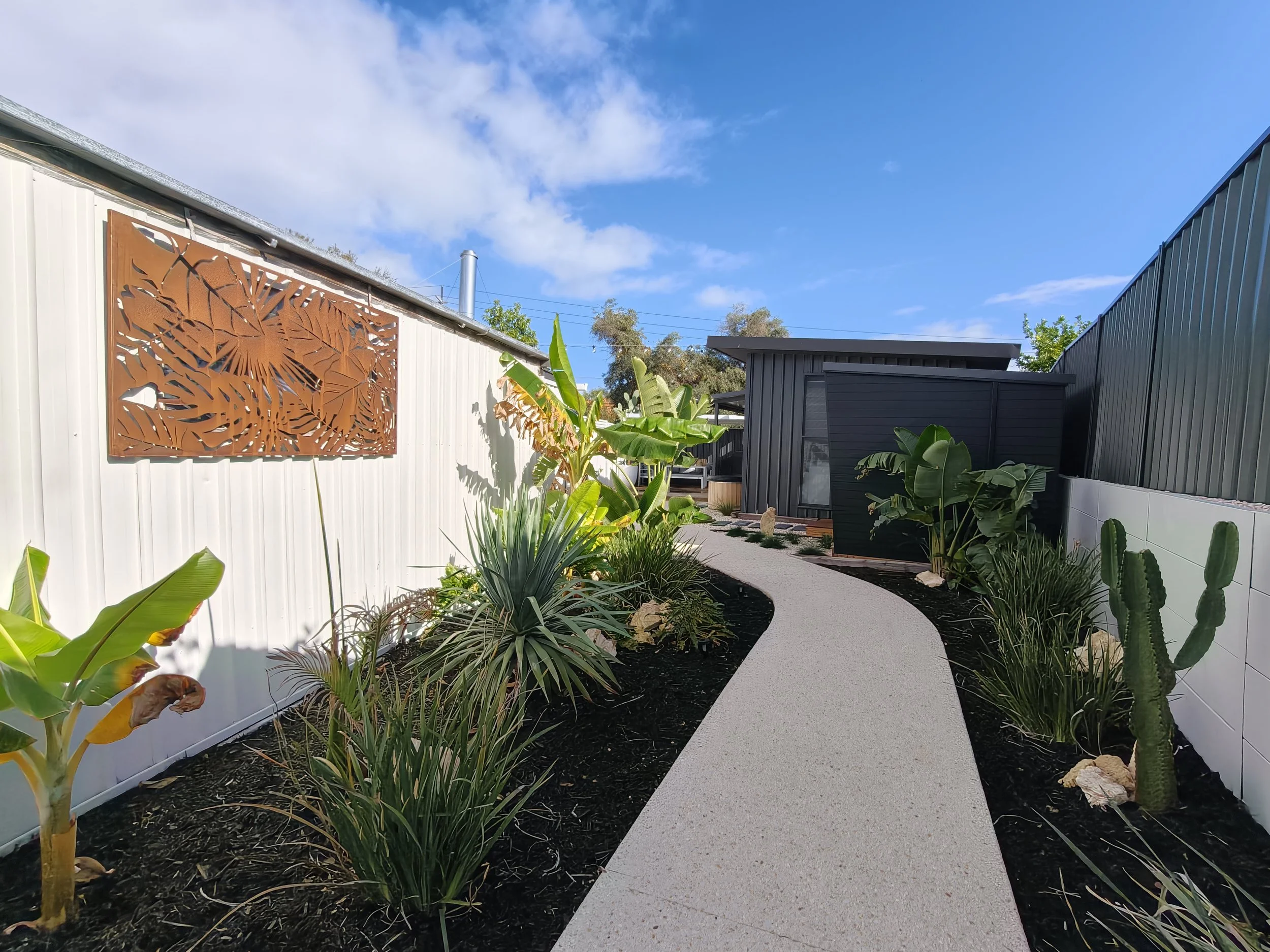 A backyard garden with a winding white path, various green plants including banana trees and cacti, a white fence with a decorative wooden panel, and a small black shed under a clear blue sky.