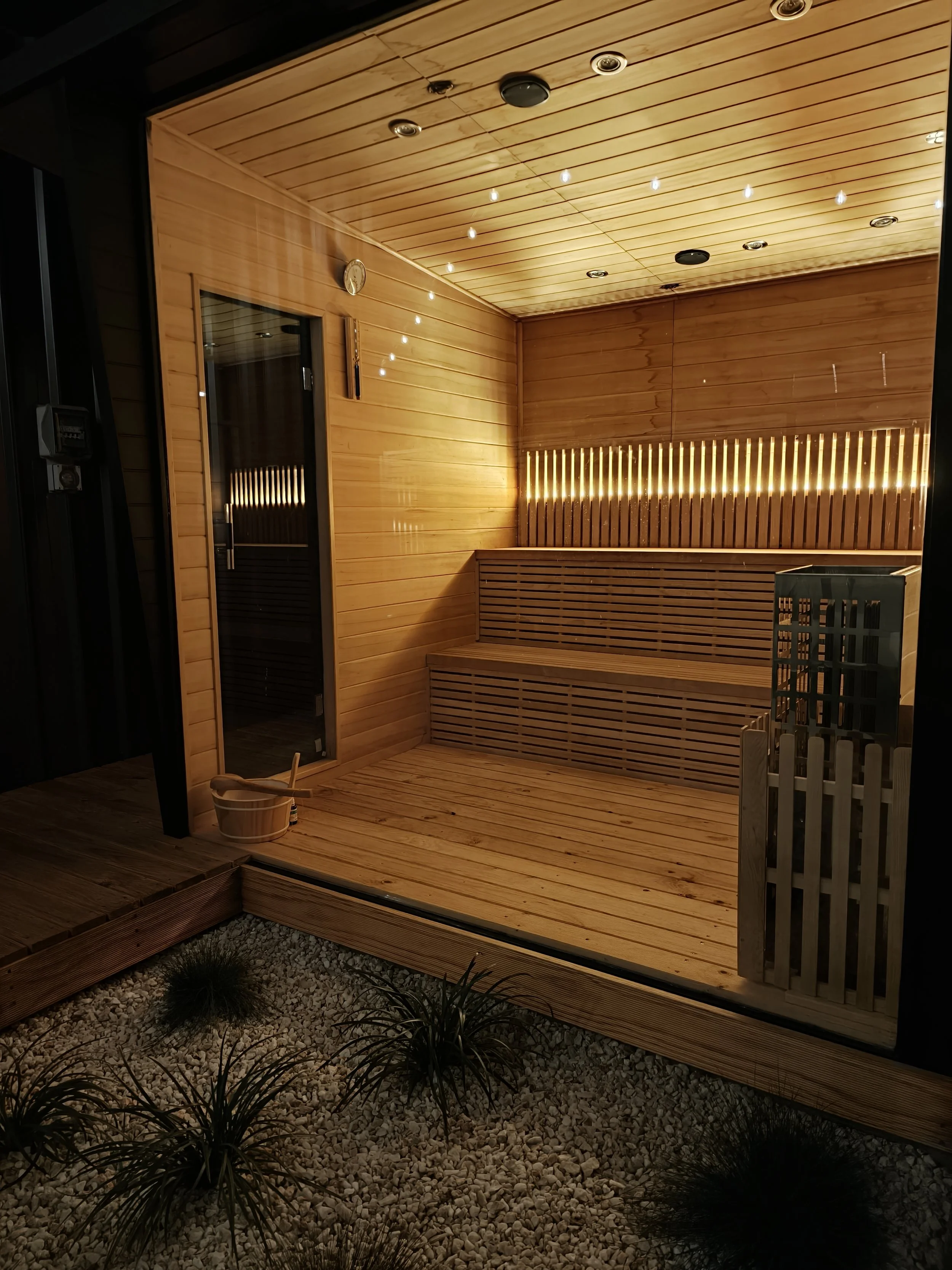 A sauna interior with wooden walls, ceiling, and benches, illuminated by warm lighting, with a small bucket and a water ladle on the floor, and decorative plants with small rocks outside.