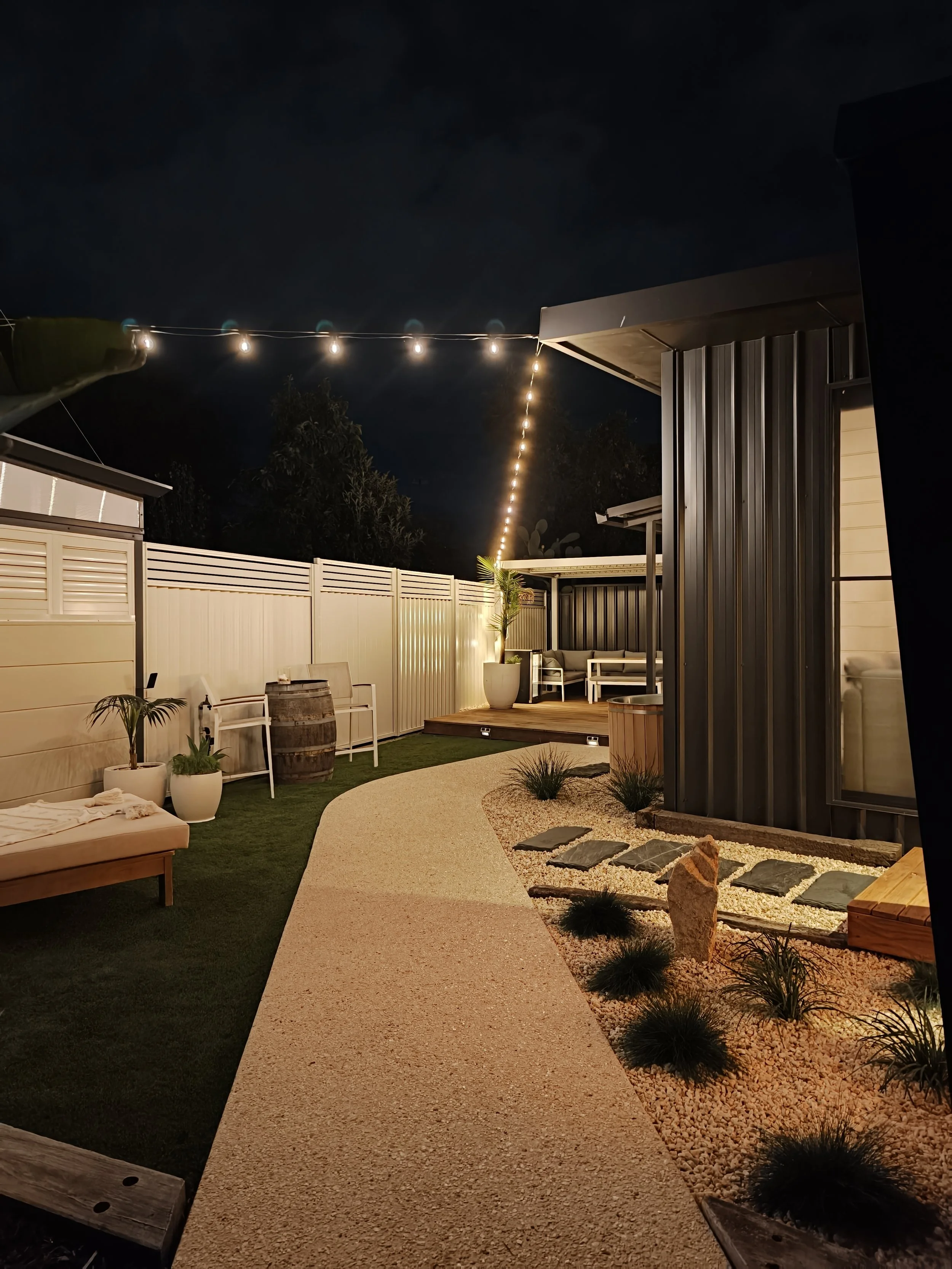 Night view of an outdoor backyard patio illuminated by string lights, featuring a pathway, potted plants, outdoor seating, and modern fencing.