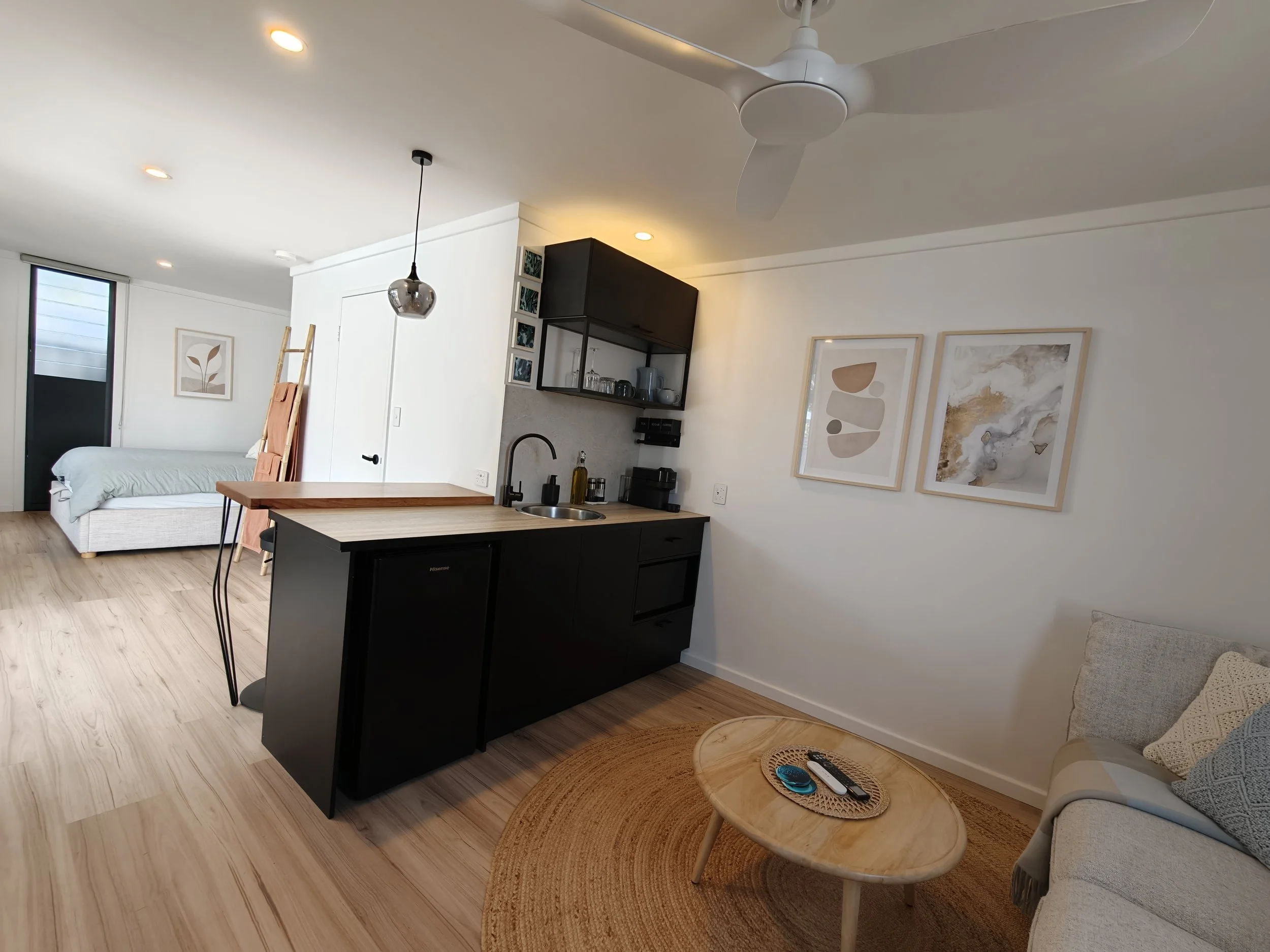 A modern living space with a white ceiling fan, recessed lighting, a hanging pendant light, a black kitchen island with a sink, and a small round wooden coffee table with a remote and phone. There is a bed with white bedding in the background, artwor
