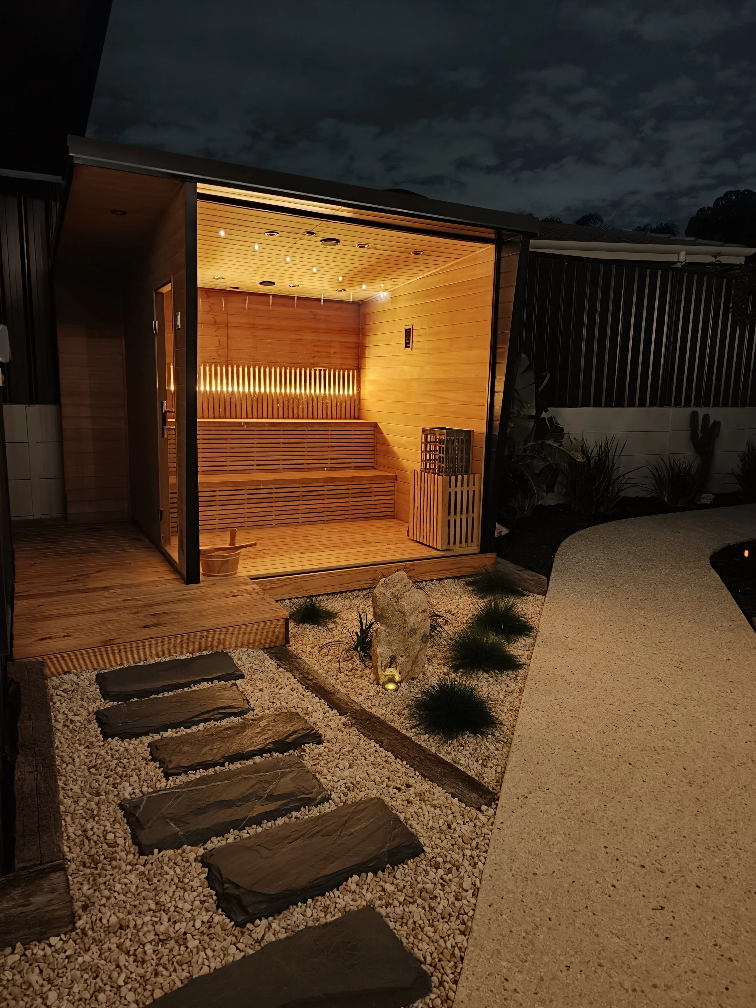 An outdoor wooden sauna illuminated at night, with a stone pathway and landscaped garden surrounding it.