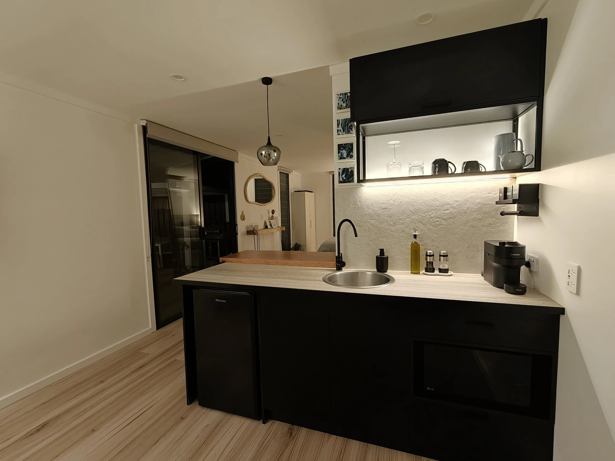 Modern kitchen with black cabinetry, light wood flooring, a dining counter, and hanging light fixture, with a reflection showing glassware and mugs.