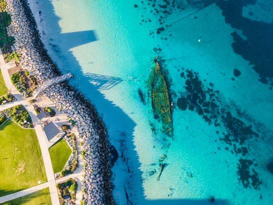 A submerged aircraft wreck on a clear blue body of water near a rocky shoreline with a park and pathway.