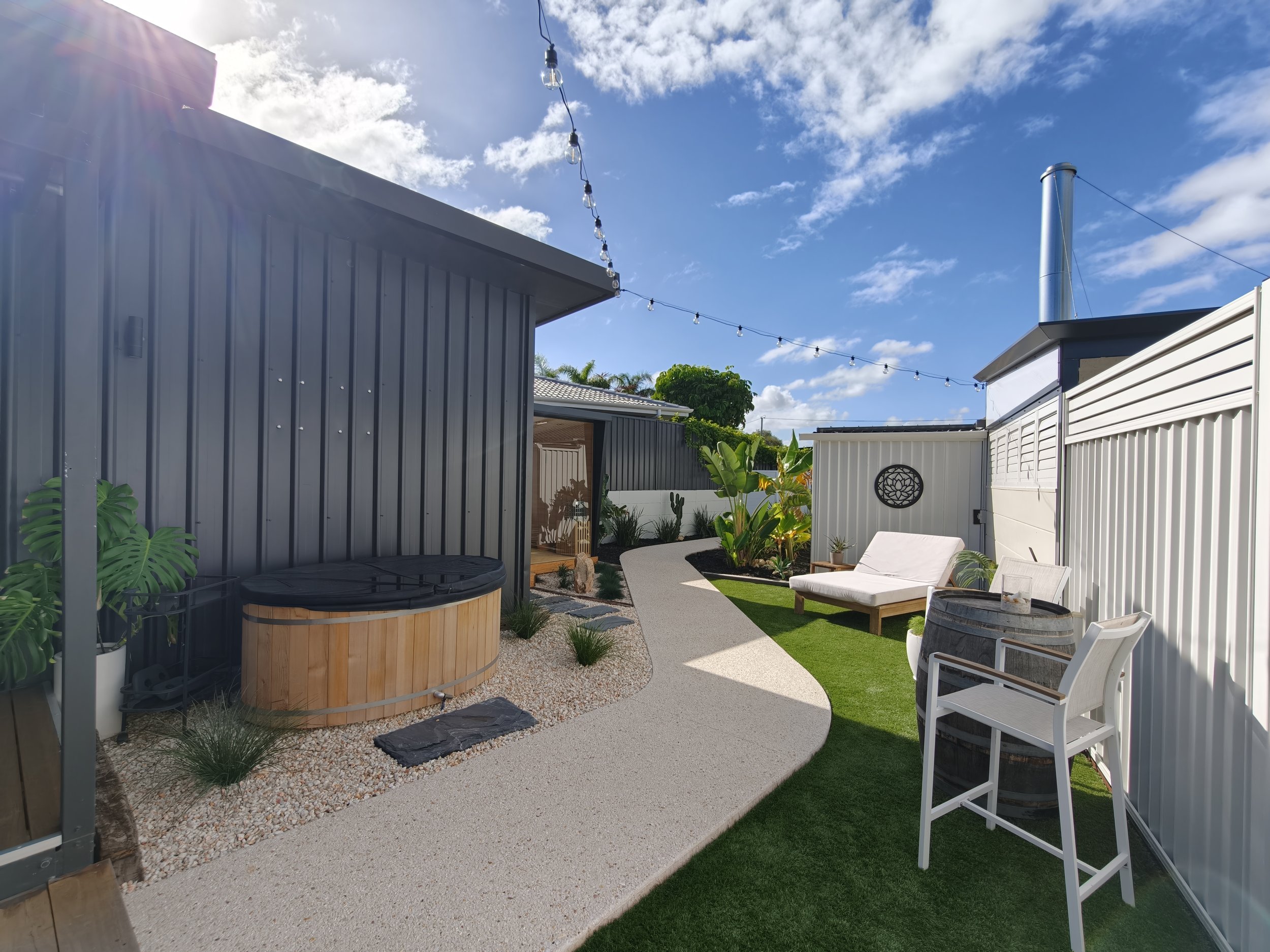 A backyard patio with a gravel and concrete pathway, lush green plants, white outdoor chairs, a hot tub, and string lights hanging above under a bright blue sky with a few clouds.