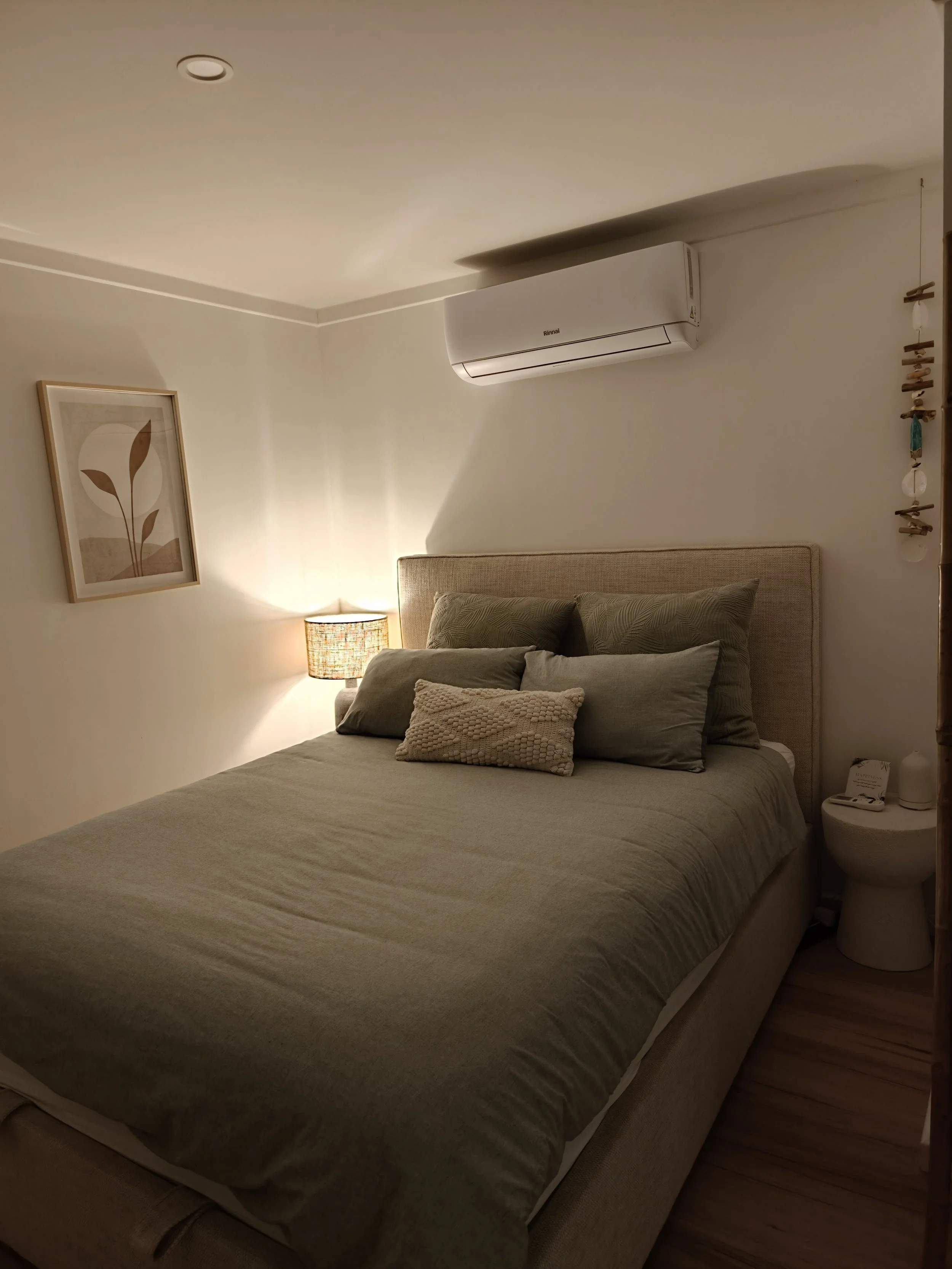 A bedroom with a neatly made bed with pillows, a nightstand with a lamp, and wall decor. An air conditioning unit is mounted on the wall.