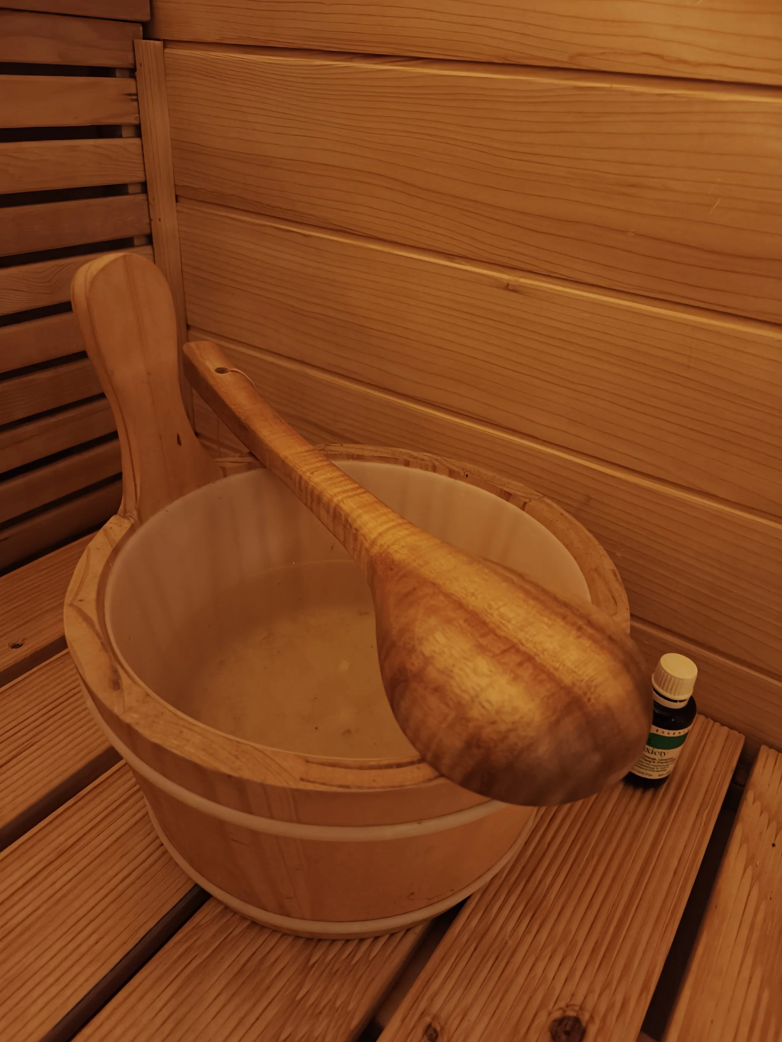 Inside a wooden sauna, a wooden bucket with a ladle and a small bottle of essential oil are placed on a wooden bench.