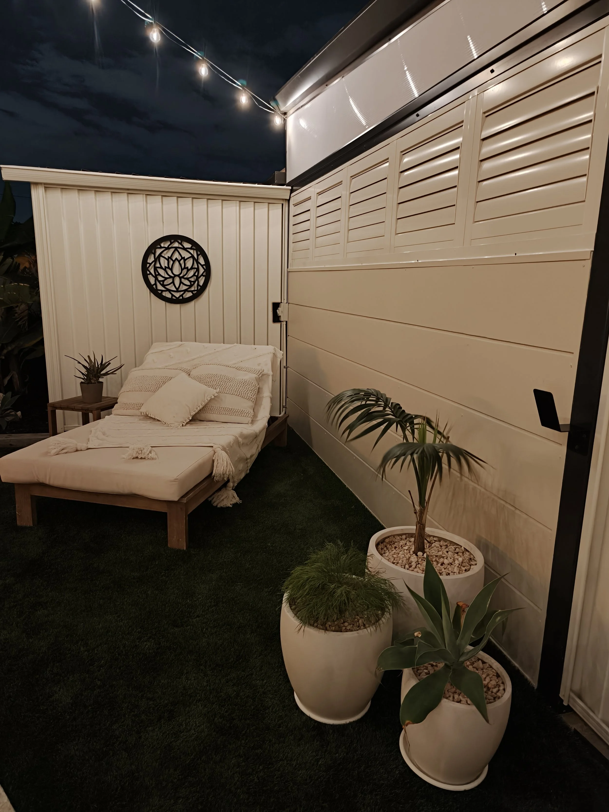 Nighttime patio scene with a bed, potted plants, string lights, and a white fence.
