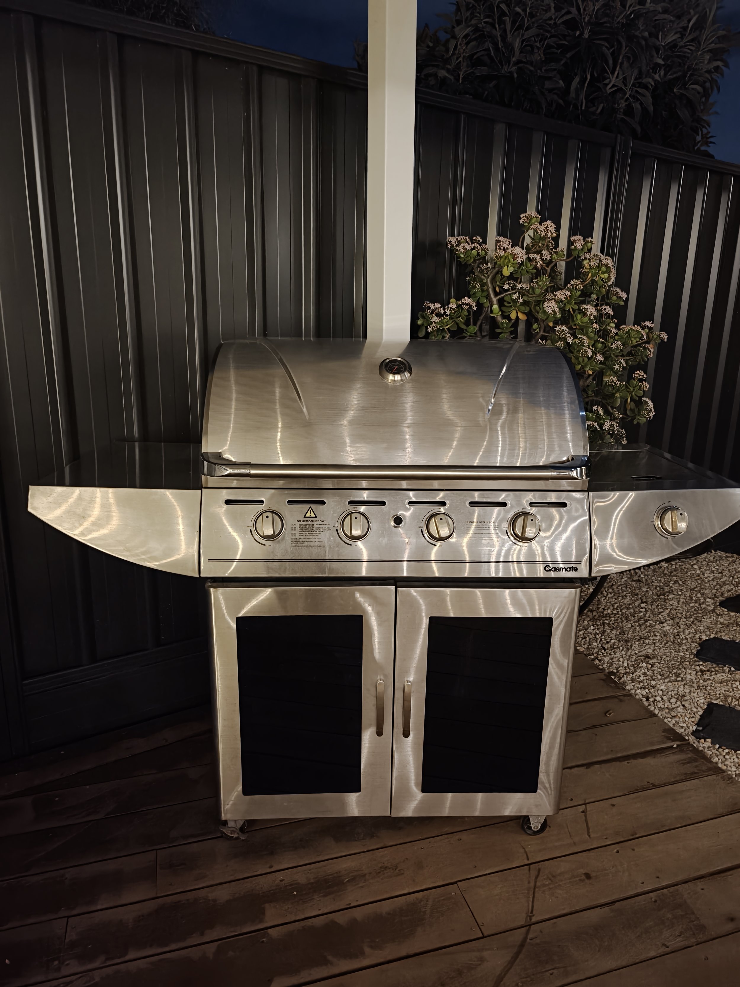 Stainless steel outdoor grill on a deck, with a black fence and decorative plants in the background.