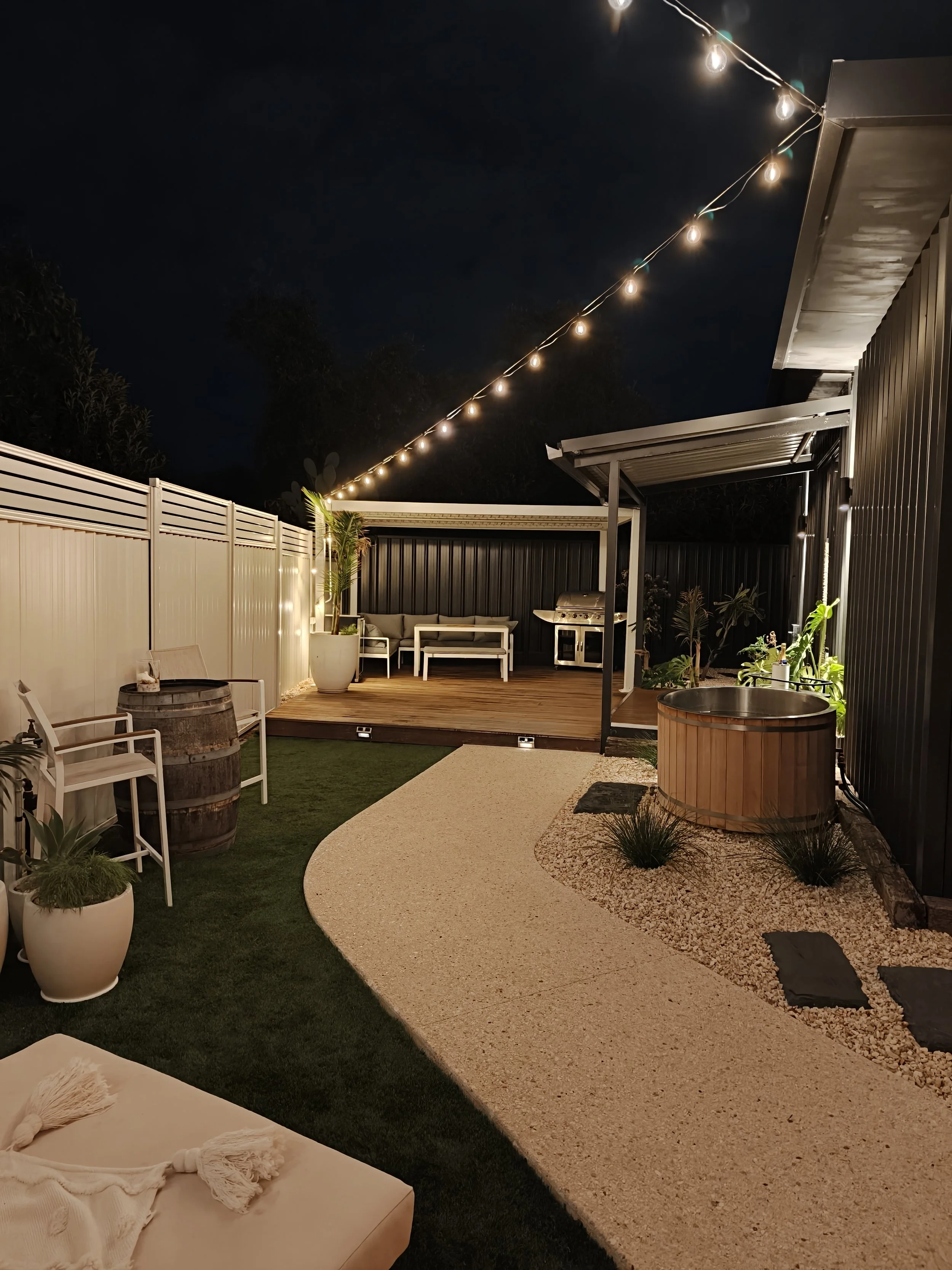 Nighttime backyard patio with string lights, a wooden deck, outdoor seating, a barbecue grill, potted plants, a hot tub, and a pathway with small stones.