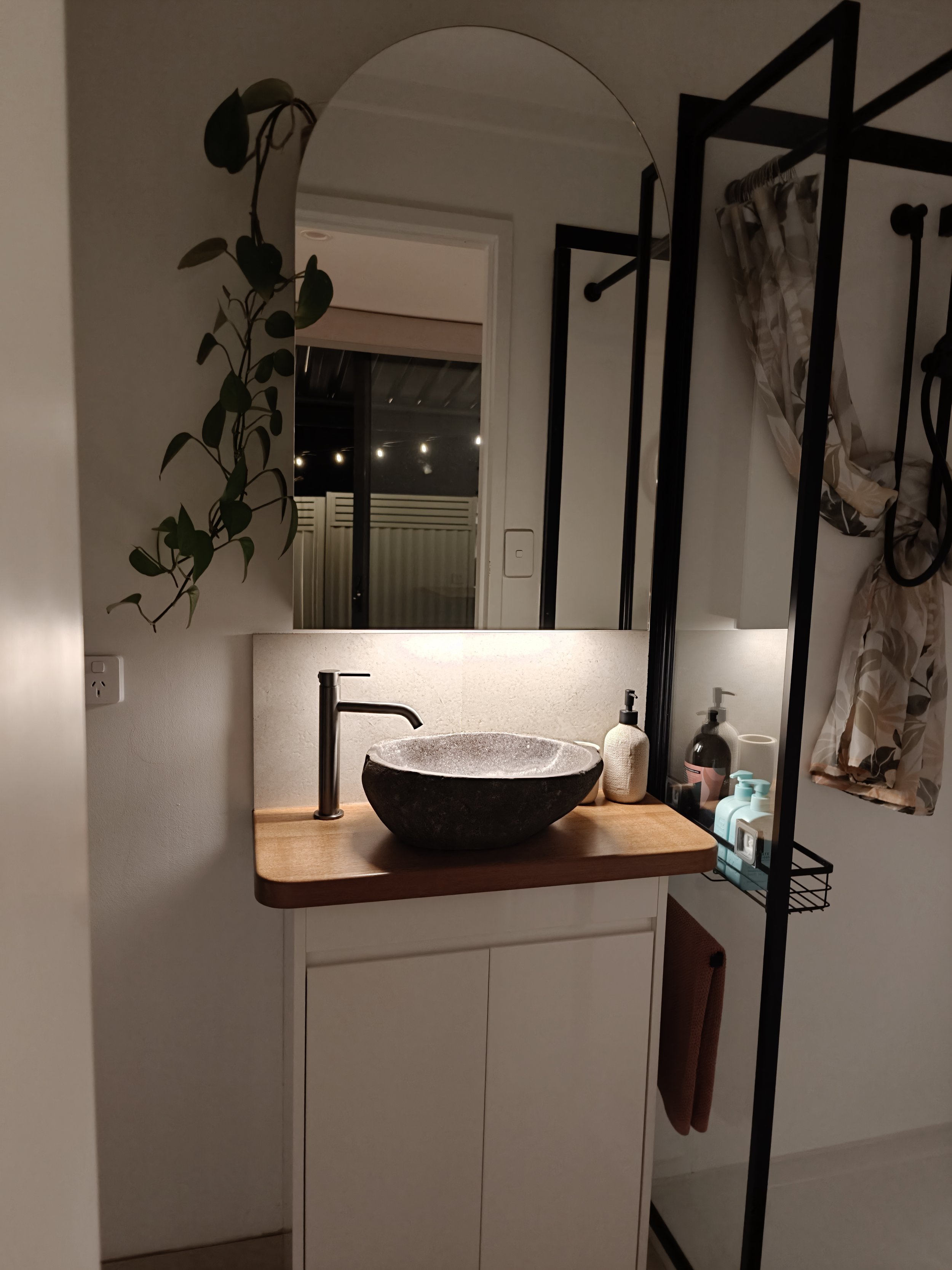 Bathroom vanity with a black stone sink, wooden countertop, and a tall mirror. There are various toiletries, a potted plant, a framed mirror, a window, a shower curtain, a towel, and a shelving unit with bottles.