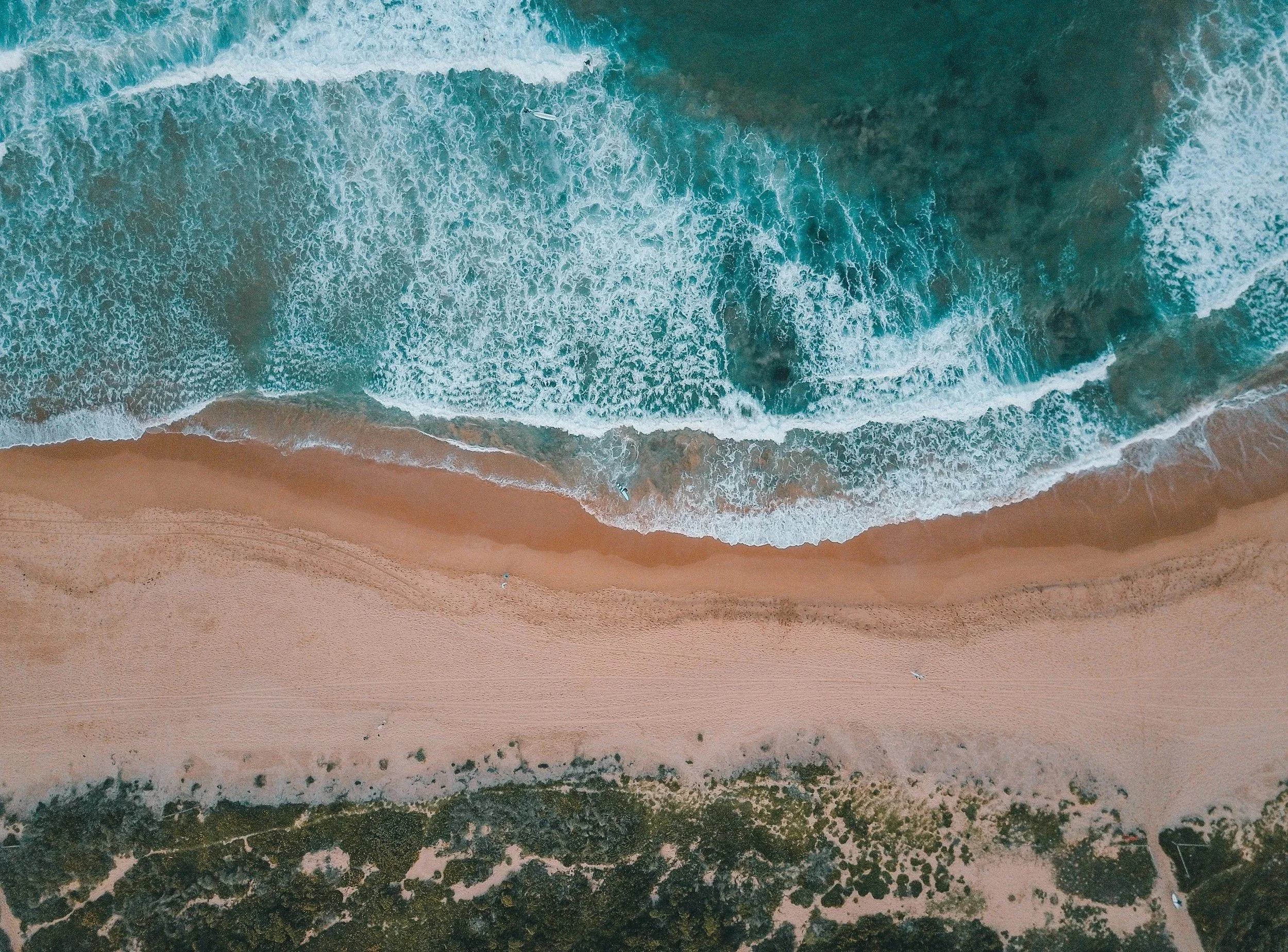 Aerial view of ocean waves crashing onto a sandy beach, with some dark patches of seaweed or rocks near the shoreline.