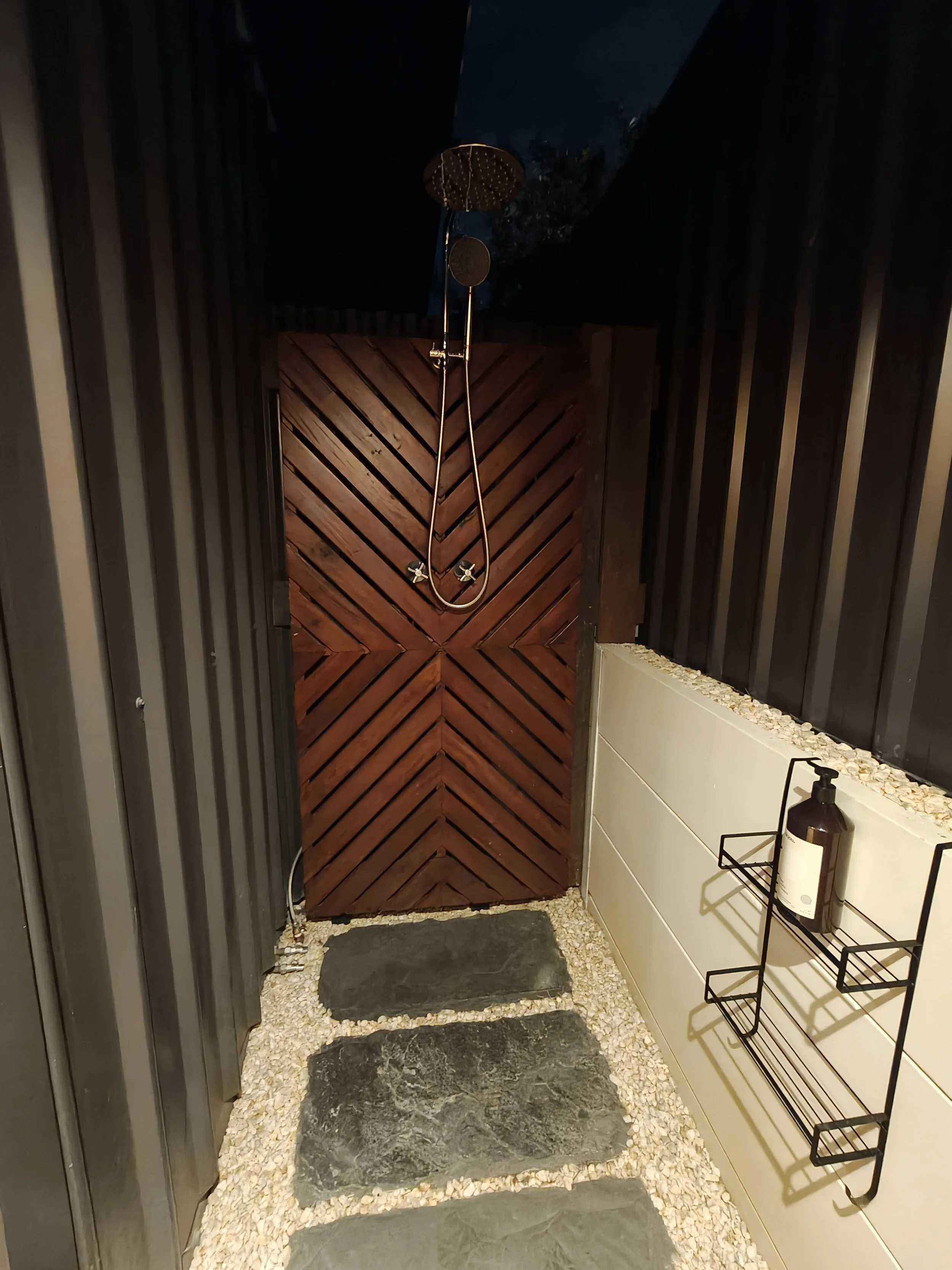 A small outdoor shower area with a wooden door at the end, black stone tiles on the floor, and white pebbles outlining the edges. There is a black metal shelf with a bottle of soap and a small container. The showerhead is mounted on the ceiling with 