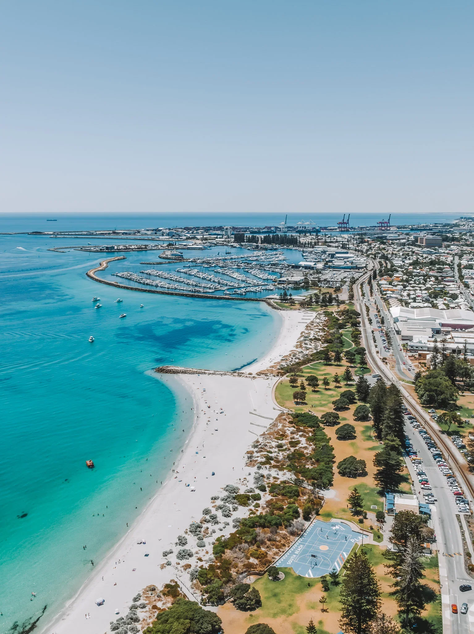 An aerial view of a coastal city with a beach, ocean, boats, a marina, trees, and a park with a basketball court.