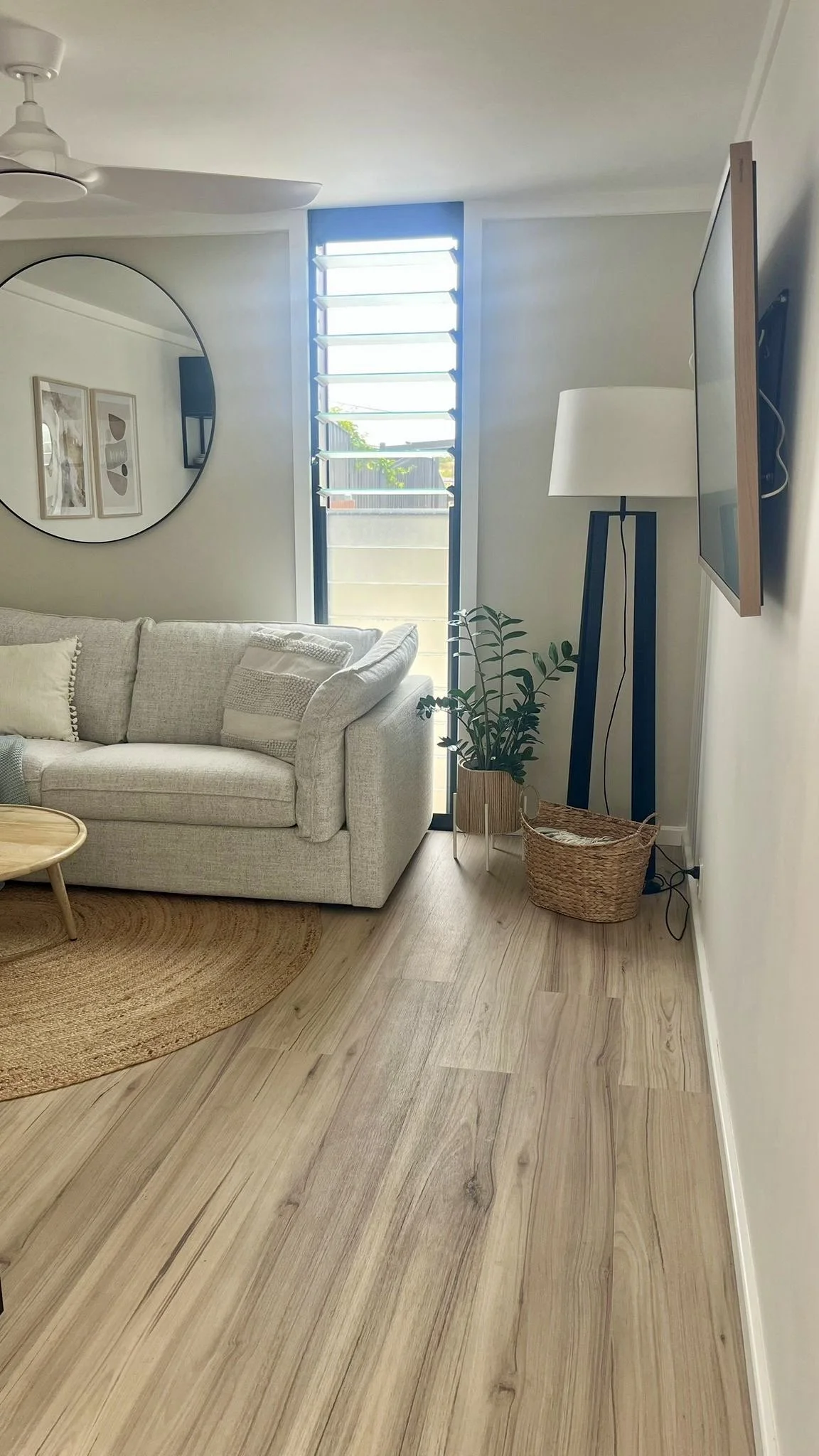 A cozy living room with a beige sofa, round wooden coffee table, round mirror on the wall, a standing lamp, a potted plant, a woven basket, and a television mounted on the wall. Natural light streams through a window with adjustable louvers.