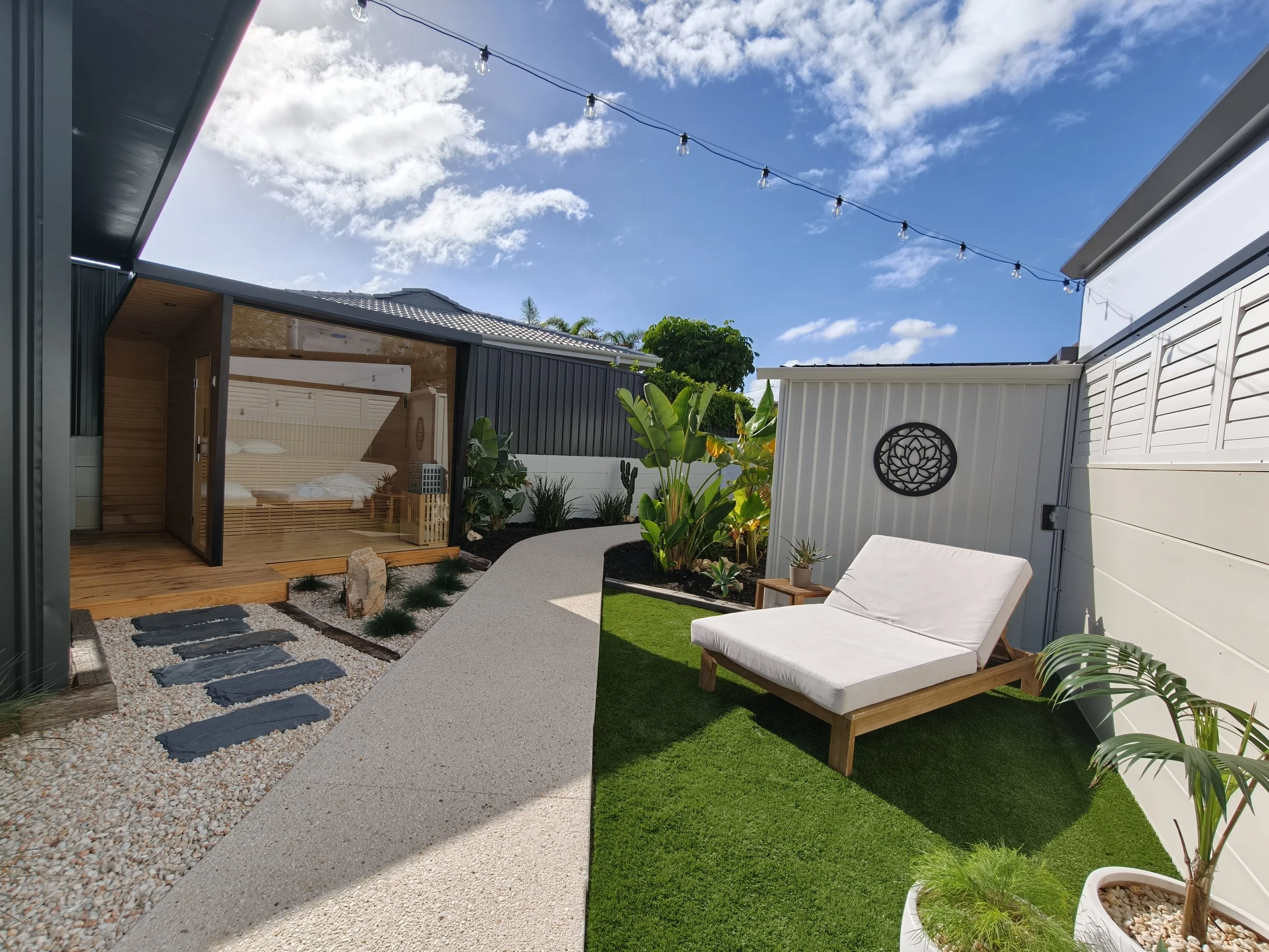 A backyard patio with a narrow pathway of black stepping stones set in white gravel, a wooden outdoor shower, a white outdoor lounge chair, lush green plants and trees, and a string of outdoor lights overhead against a blue sky with some clouds.