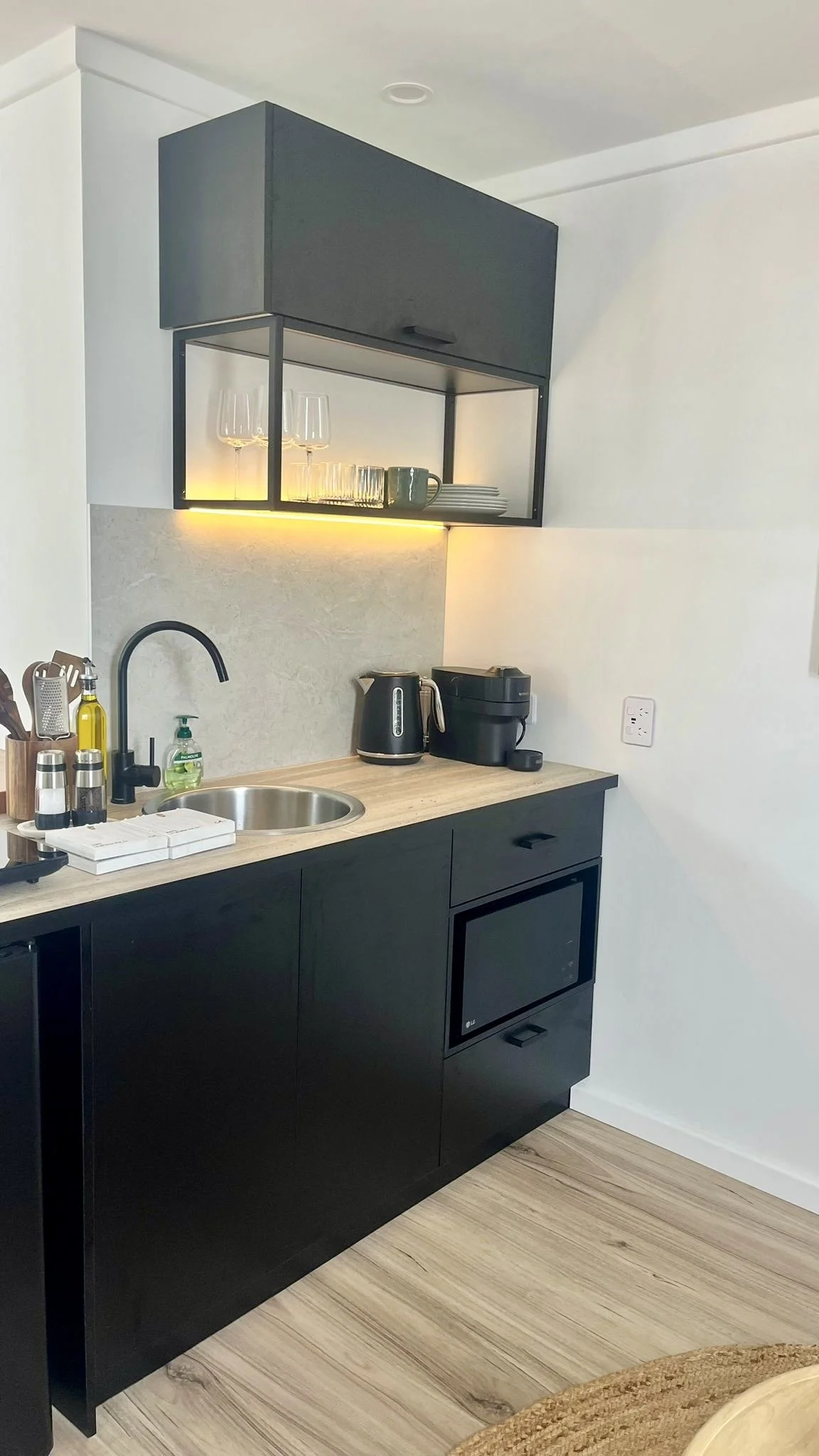 A modern kitchen corner with black cabinets, a wooden countertop, a small round sink with a black faucet, and kitchen appliances like a kettle and rice cooker. Open shelving with glasses, plates, and cups, under warm lighting.