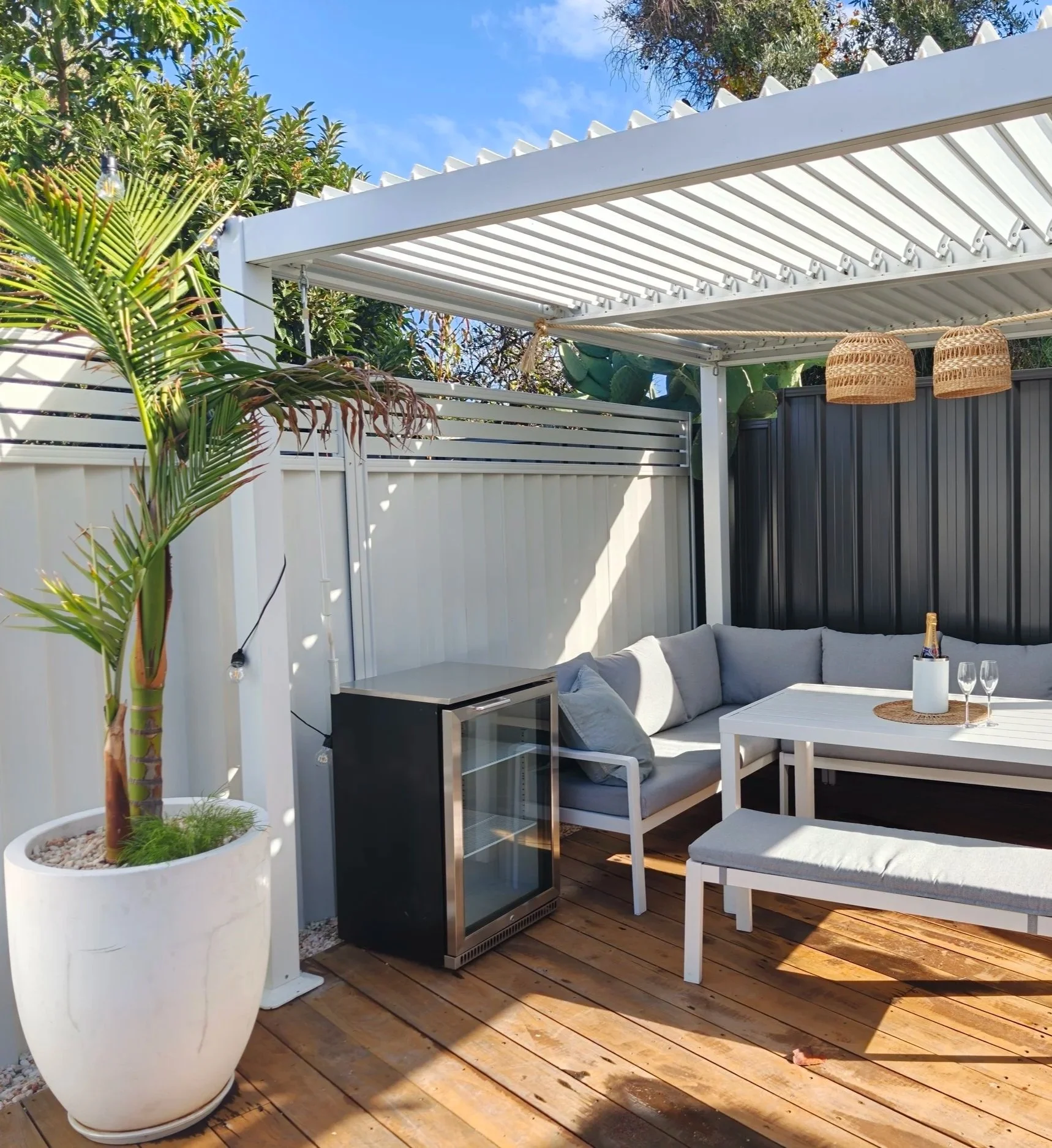 Outdoor patio with white and black furniture, potted plant, and wooden deck flooring under a white pergola with hanging wicker lamps and string lights.