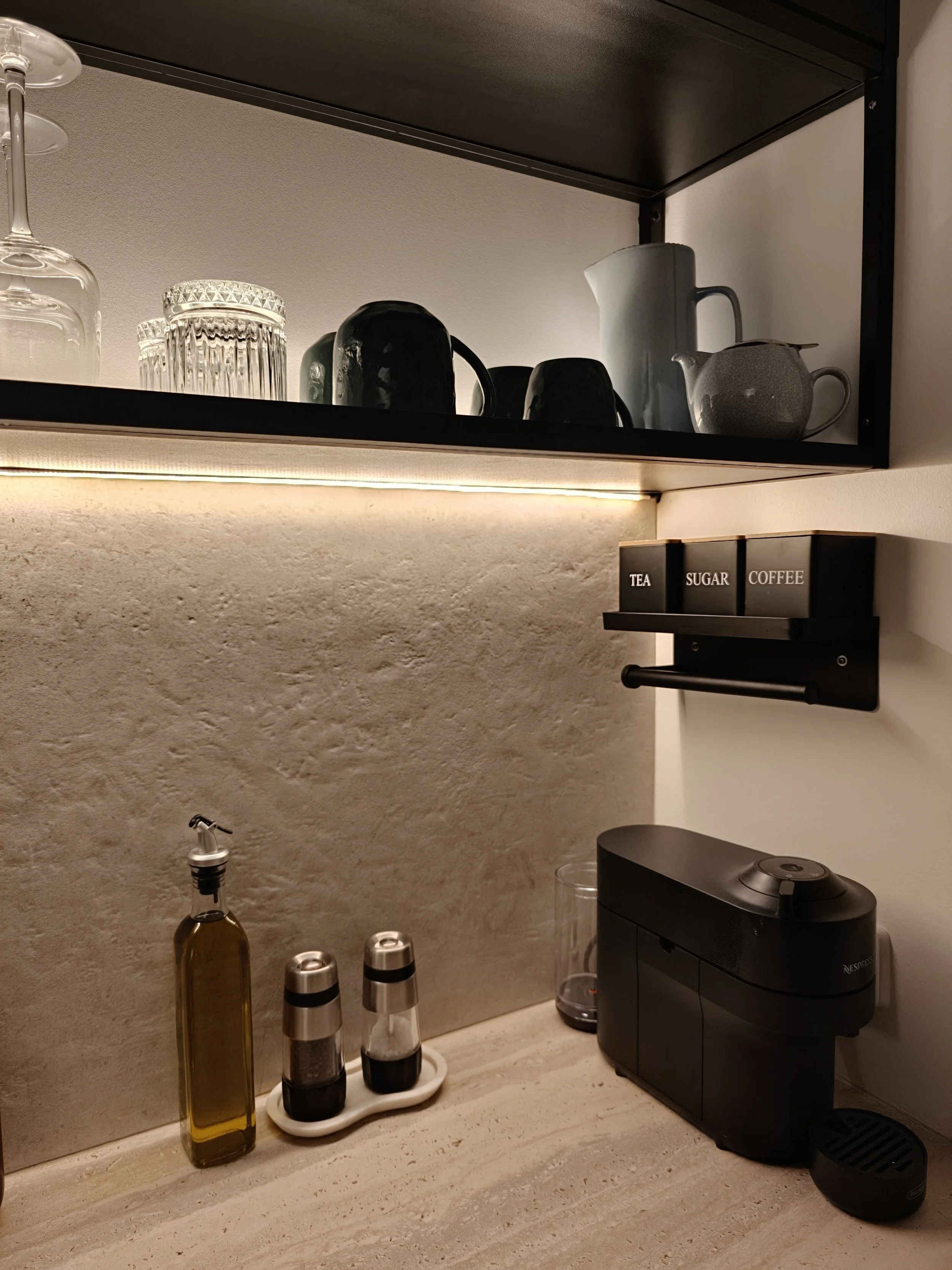 Kitchen shelf with glasses, cups, teapot, pitchers, and coffee machine, with a small organized spice or condiment rack labeled "TEA," "SUGAR," and "COFFEE".