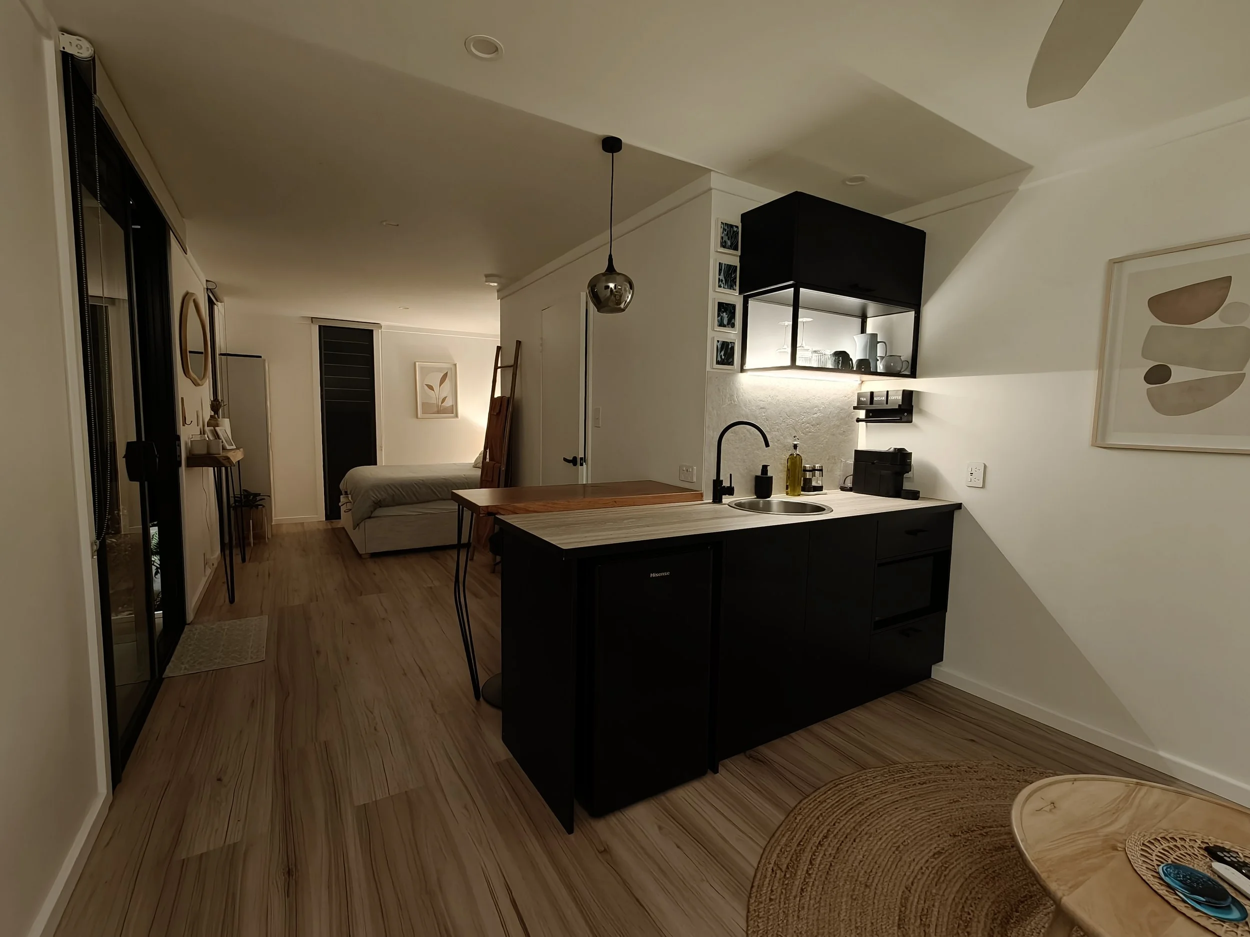 A modern open-concept living space with a kitchen area featuring black cabinetry, a wooden countertop, and a sink with black fixtures. There is a partial view of a bedroom with a bed and a decorative framed art piece on the wall. The flooring is wood