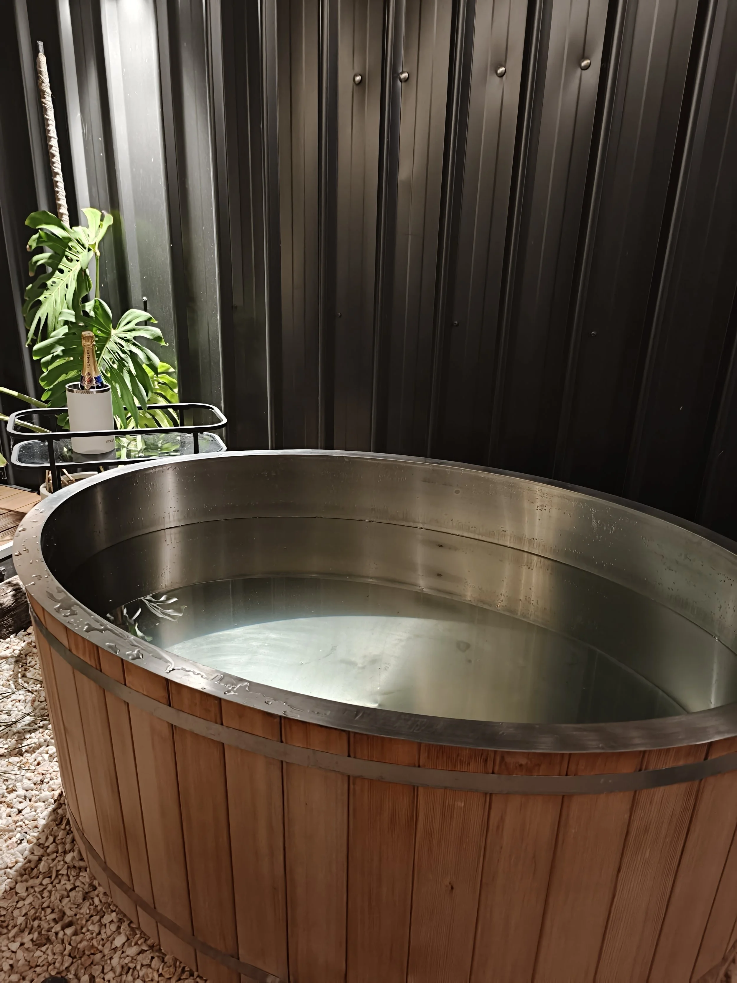 A round hot tub with wooden siding and metal trim filled with water, placed on a gravel surface next to black metal siding, with a potted Monstera plant and a bottle of champagne on nearby furniture.