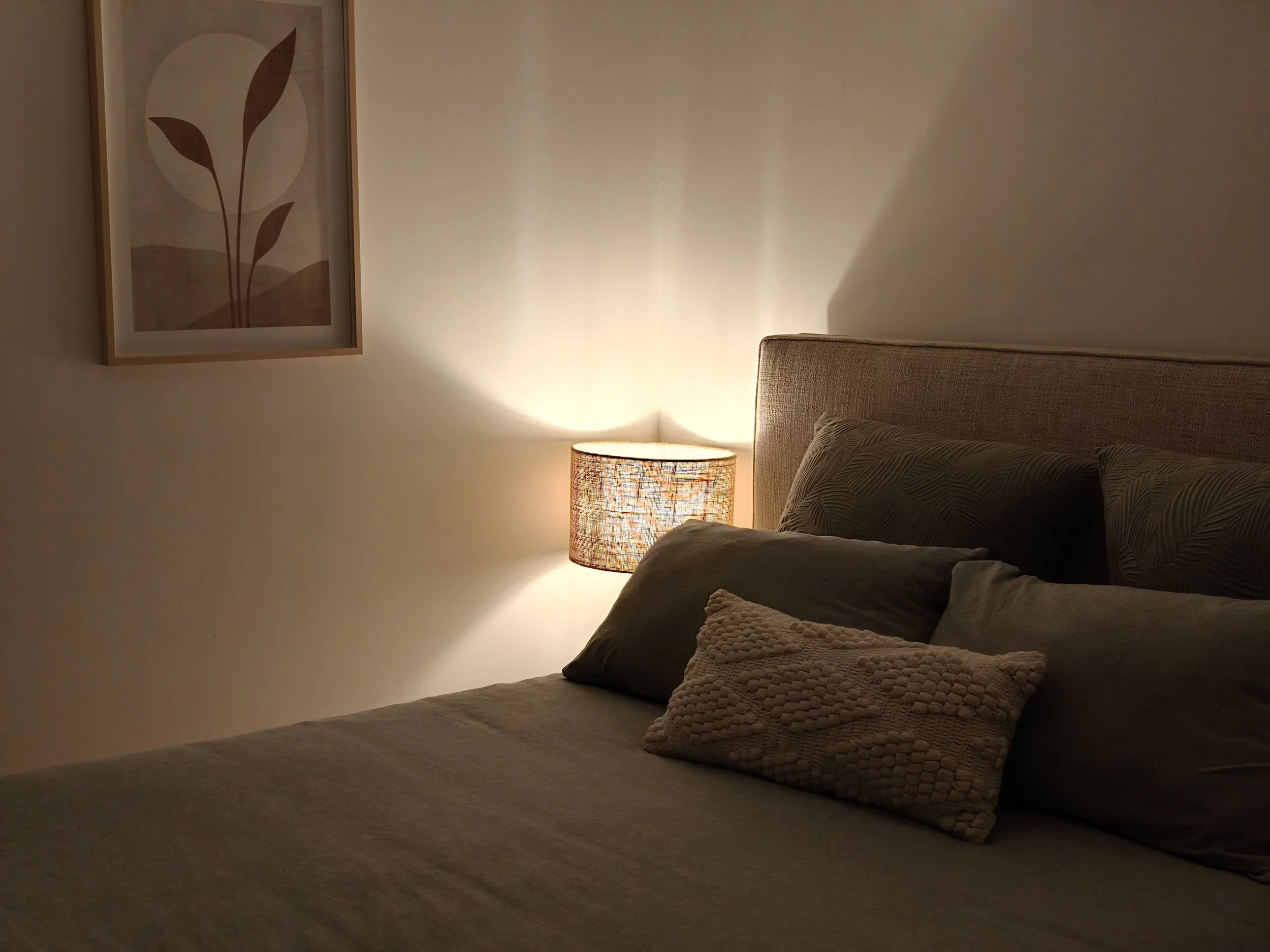 A bedroom corner with a beige bed, stacked pillows, and a lamp on a nightstand. A framed artwork with a plant design is hanging on the wall above the bed, and it is softly lit with warm lighting.