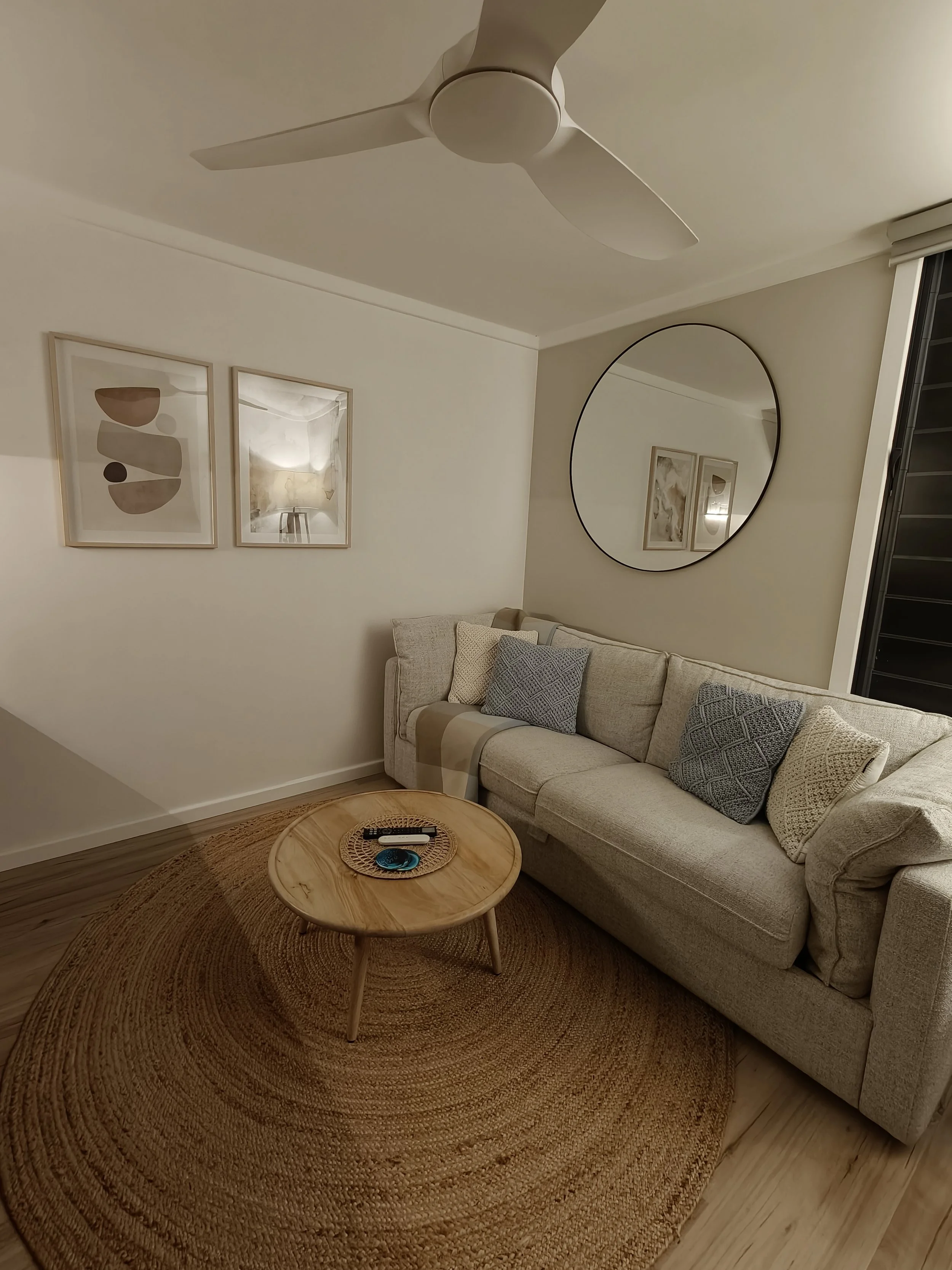 Living room with beige sofa, patterned throw pillows, round woven rug, wooden coffee table with remote controls, art prints on the wall, large circular mirror, ceiling fan, window with blinds.