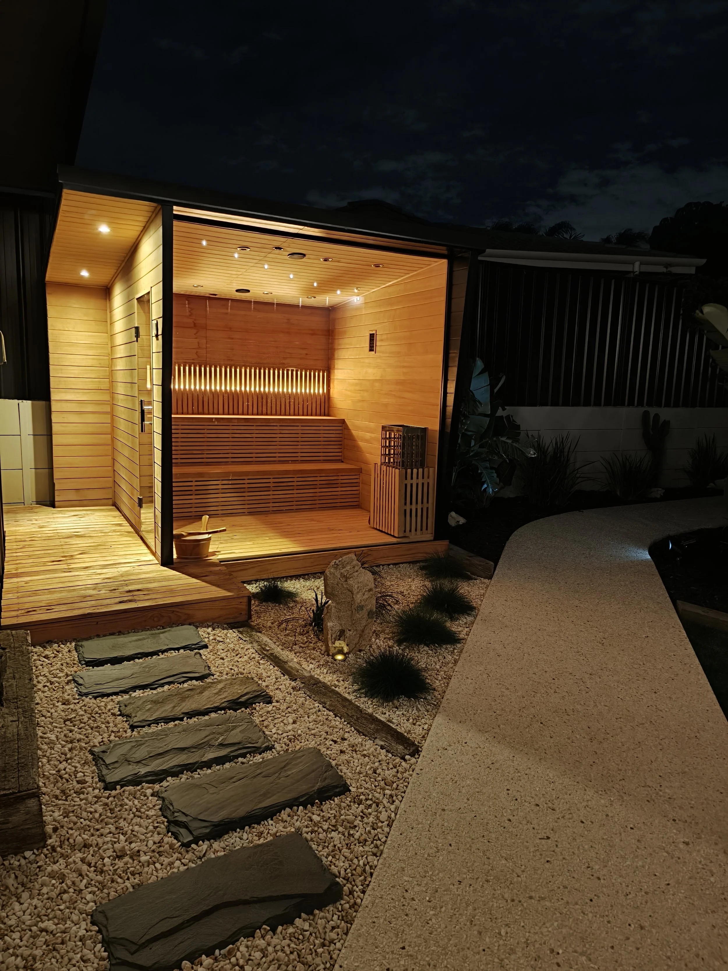 An outdoor wooden sauna illuminated at night, with a stone pathway leading to it, surrounded by decorative rocks and plants.
