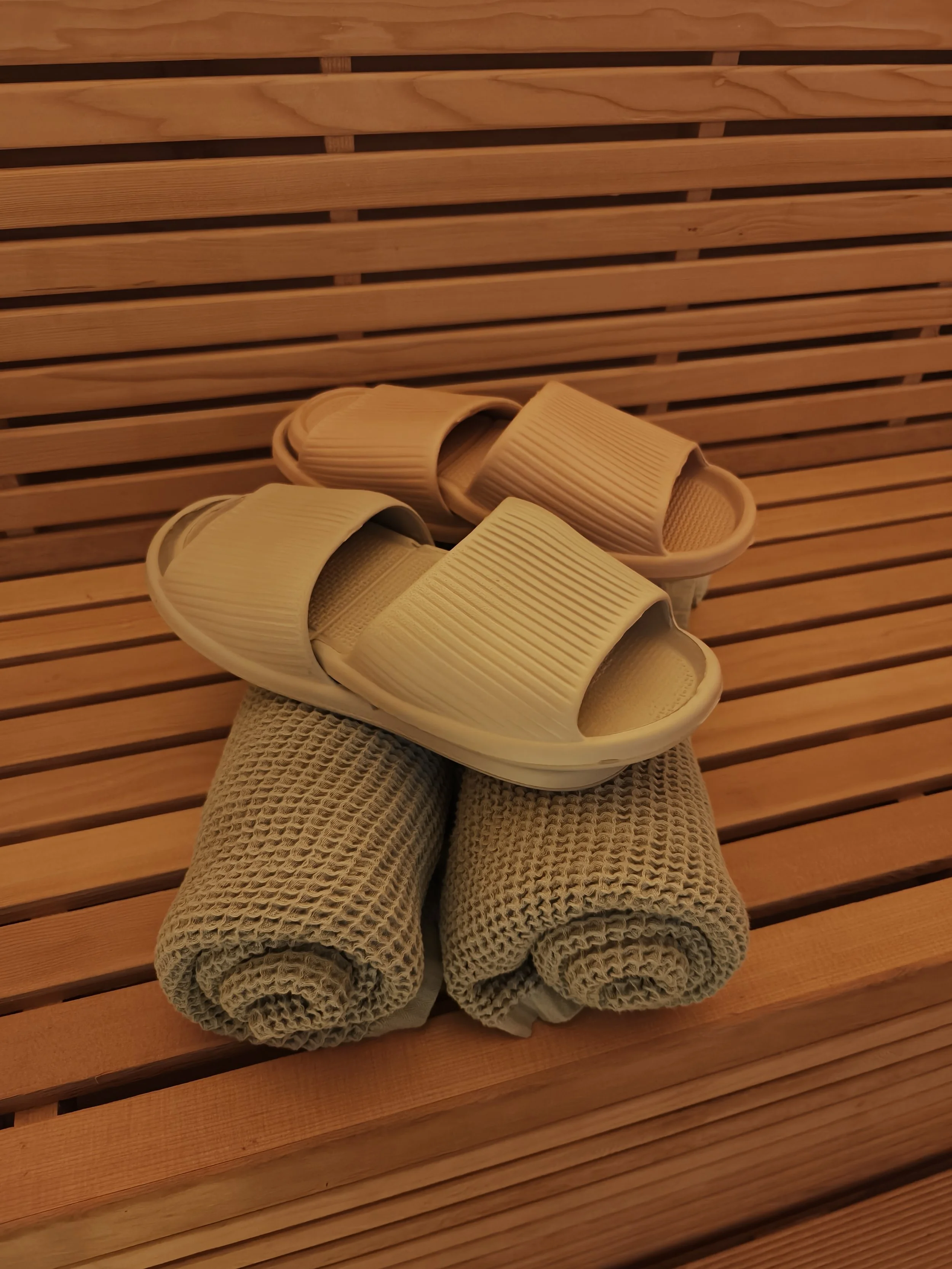 A pair of beige slippers and two rolled towels on a wooden slatted surface, likely a sauna or spa setting.