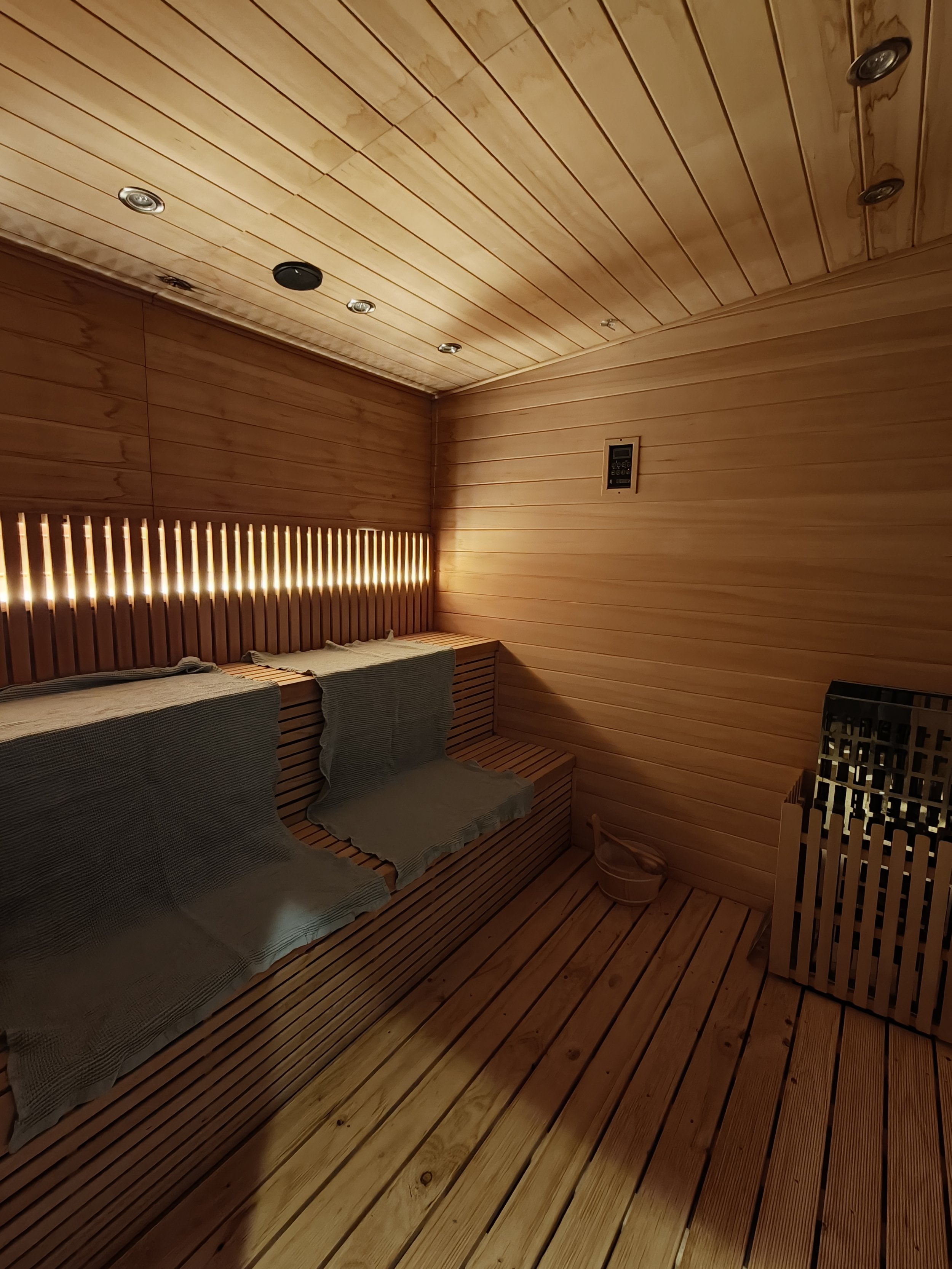 Interior view of a wooden sauna room with slatted walls, ceiling, and bench, featuring built-in lighting and a bucket in the corner.