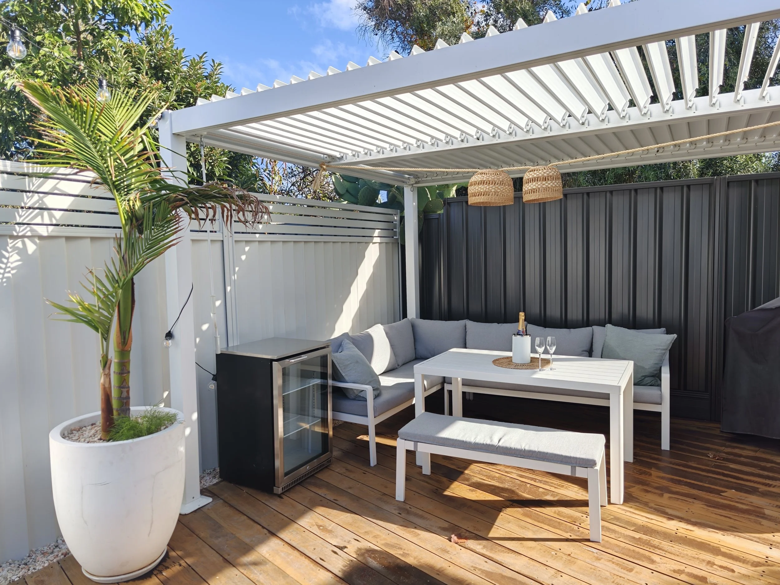A backyard patio with a white pergola, black and white fence, gray couch, small white table, large potted plant, and two wicker hanging light fixtures, with trees and blue sky in the background.